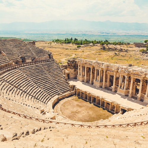 Sinaunang amphitheater na bato na may nakabatay na mga upuan at may entablado na may mga haligi, na matatagpuan sa isang malawak at bukas na kalupaan sa ilalim ng malinaw na kalangitan.