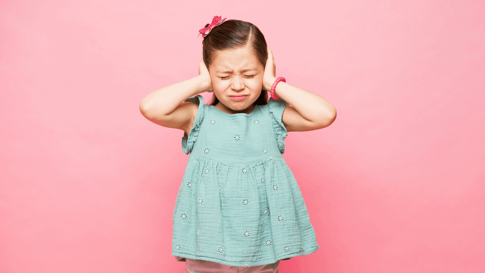 A young girl standing and covering her ears, looking stressed during a meltdown, needing something to block the background noise.