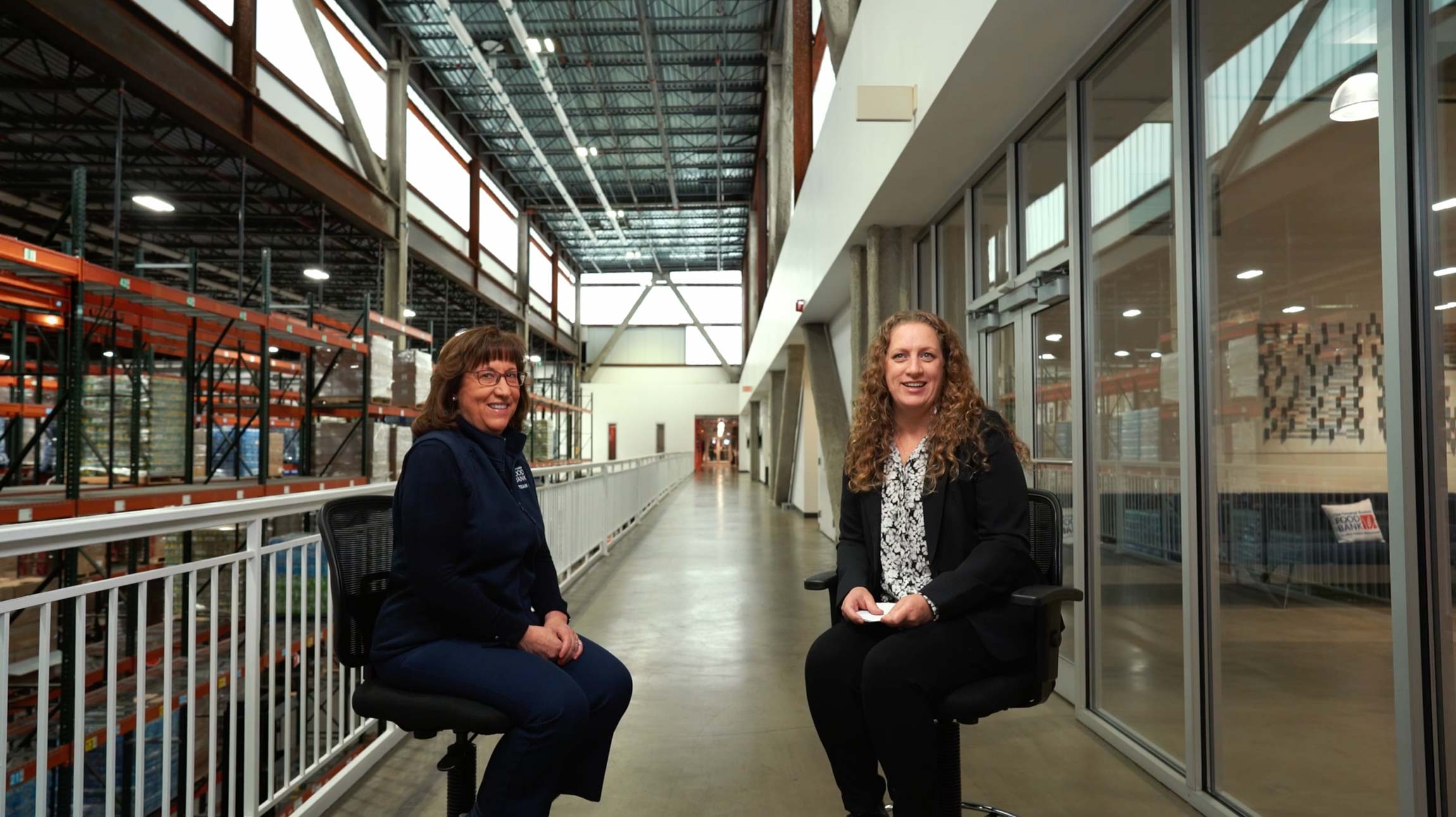 Two women sit facing the camera on office chairs in a wide hallway with warehouse shelving on the left and glass-walled rooms on the right.