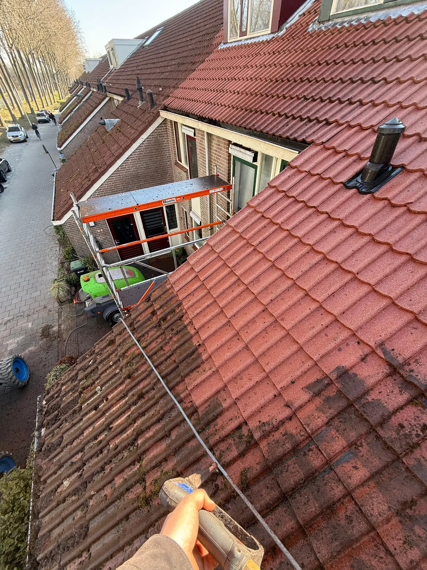 Medewerker reinigt vanaf het dak met een hogedruklans de pannen van een rij woningen; links de nog bemoste pannen, rechts het schoongespoelde deel, met een rolsteiger op straat