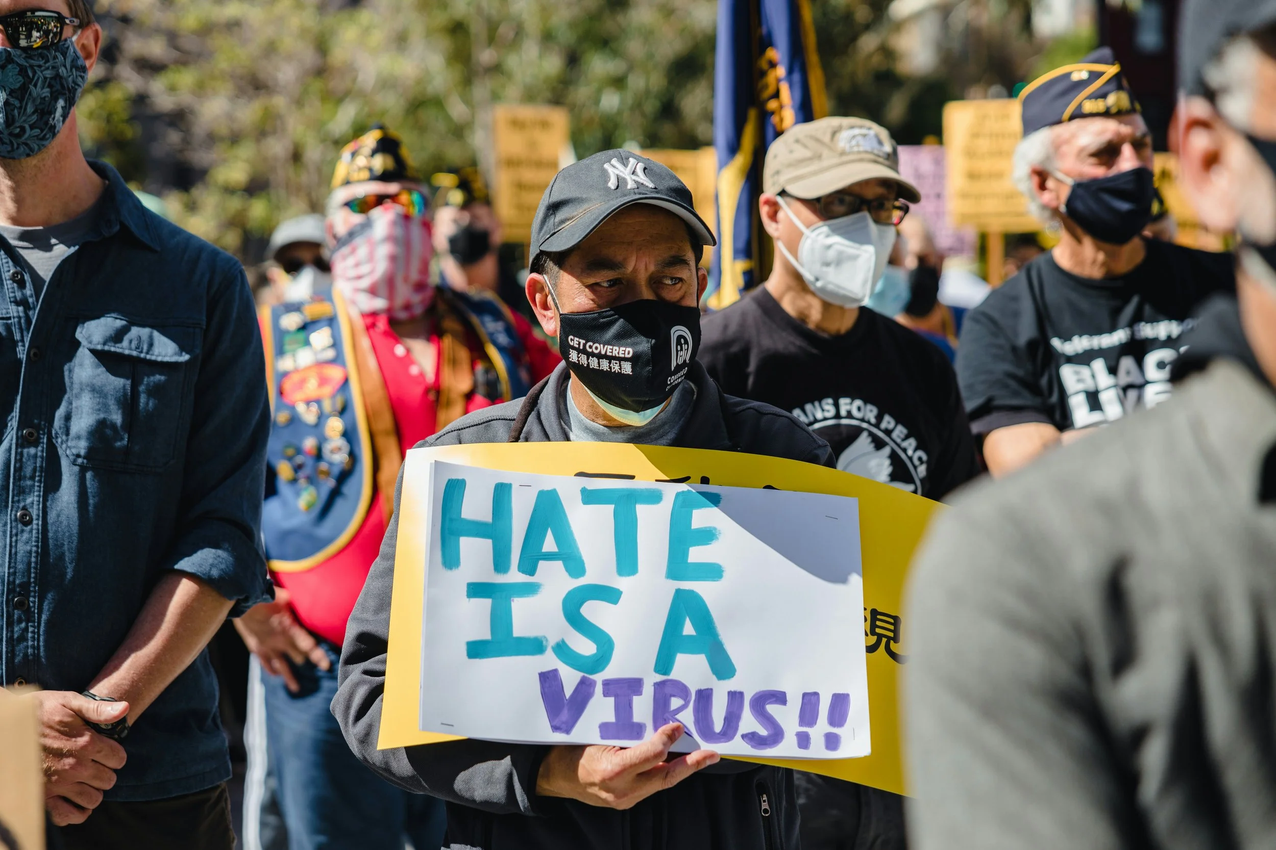 Um grupo de pessoas usando máscaras faciais e bonés participa de um protesto, segurando um cartaz colorido com os dizeres "O ódio é um vírus", tendo ao fundo árvores e luz solar intensa.
