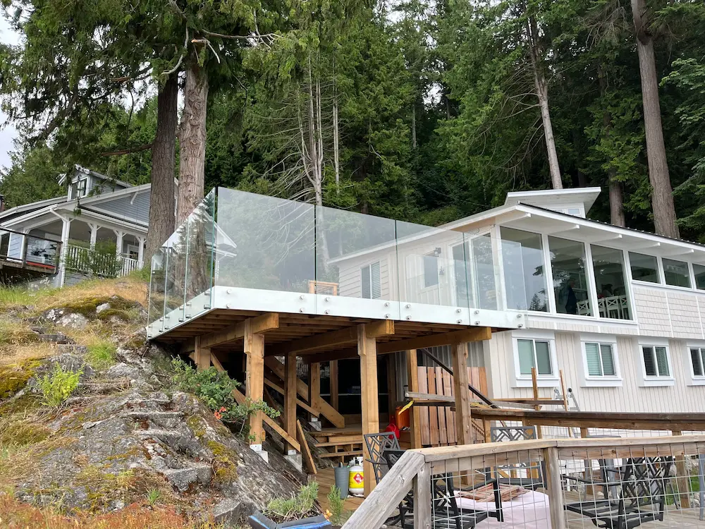 Frameless glass deck railing on a lakefront cabin built into a rocky hillside, providing clear views of the surrounding forest and waterfront.