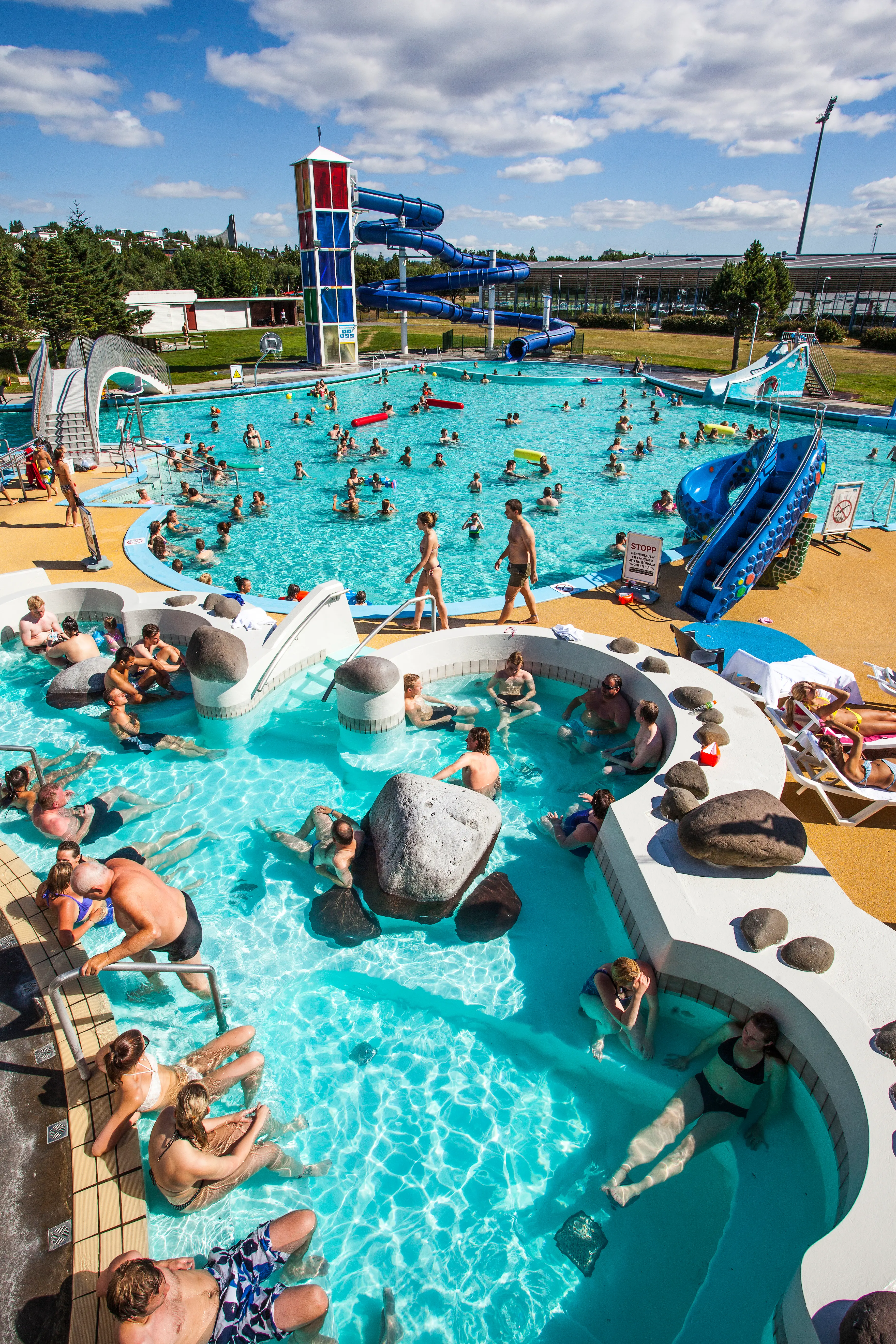 Colorful outdoor scene at Laugardalslaug, Reykjavík’s largest geothermal swimming pool, on a sunny day. Families and swimmers enjoy various heated pools, hot tubs, waterslides, and sun loungers, with the main stadium structure and blue sky in the background.