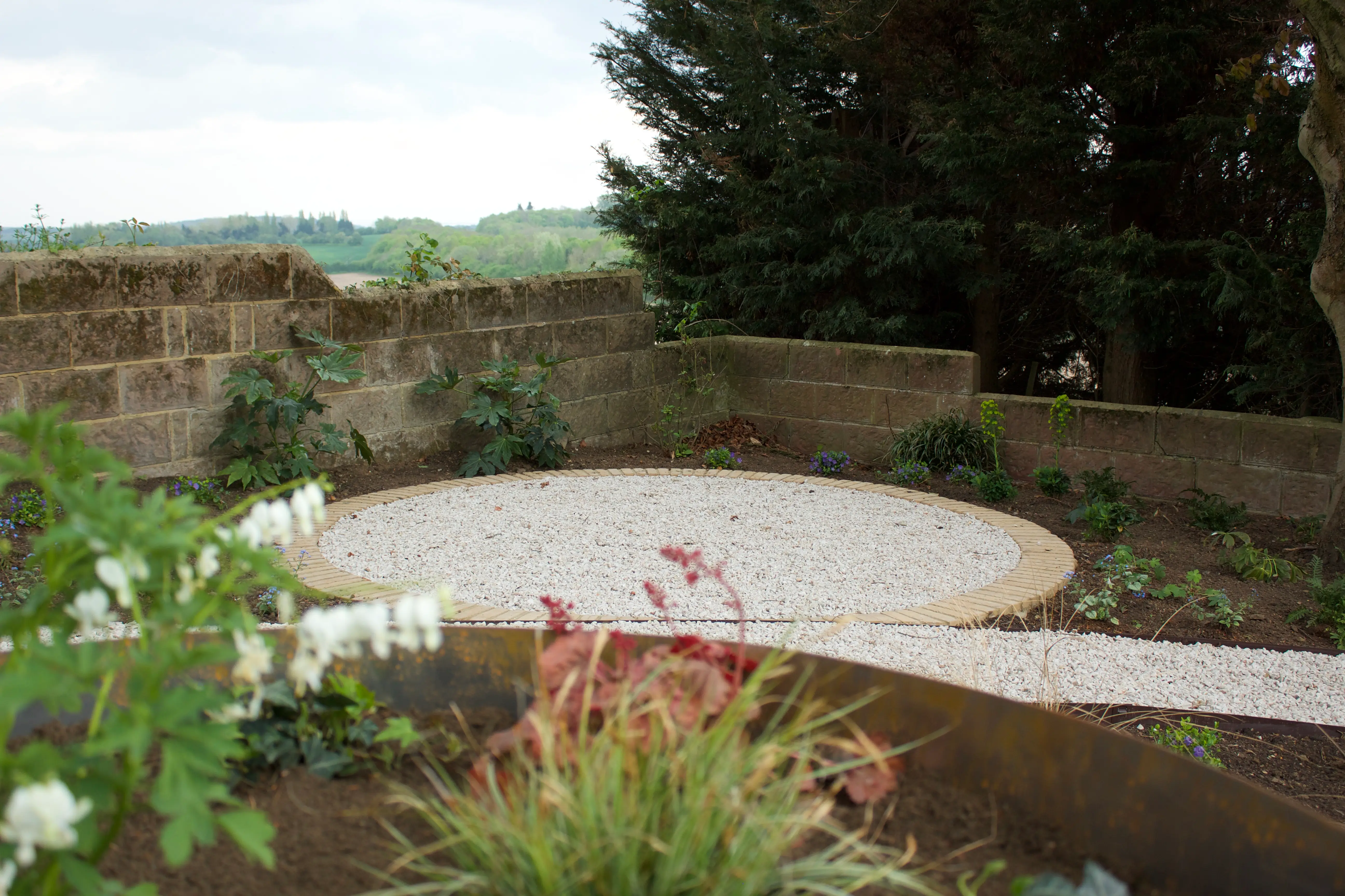 A circular stone path surrounded by greenery and flowers, set against a backdrop of trees and a cloudy sky.