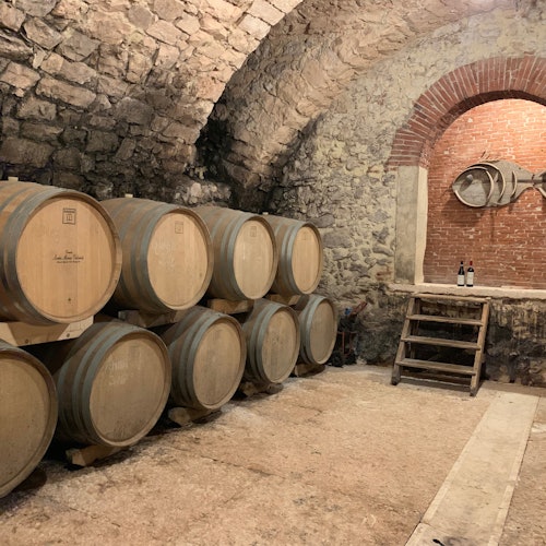 Bodega de piedra con barriles de madera apilados, una pequeña escalera que conduce a un arco de ladrillo y dos botellas de vino en una mesa.