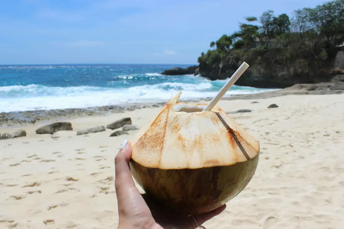 Hand holding a fresh coconut on a sunny Bali beach, symbolizing relaxation and hydration after yoga practice.