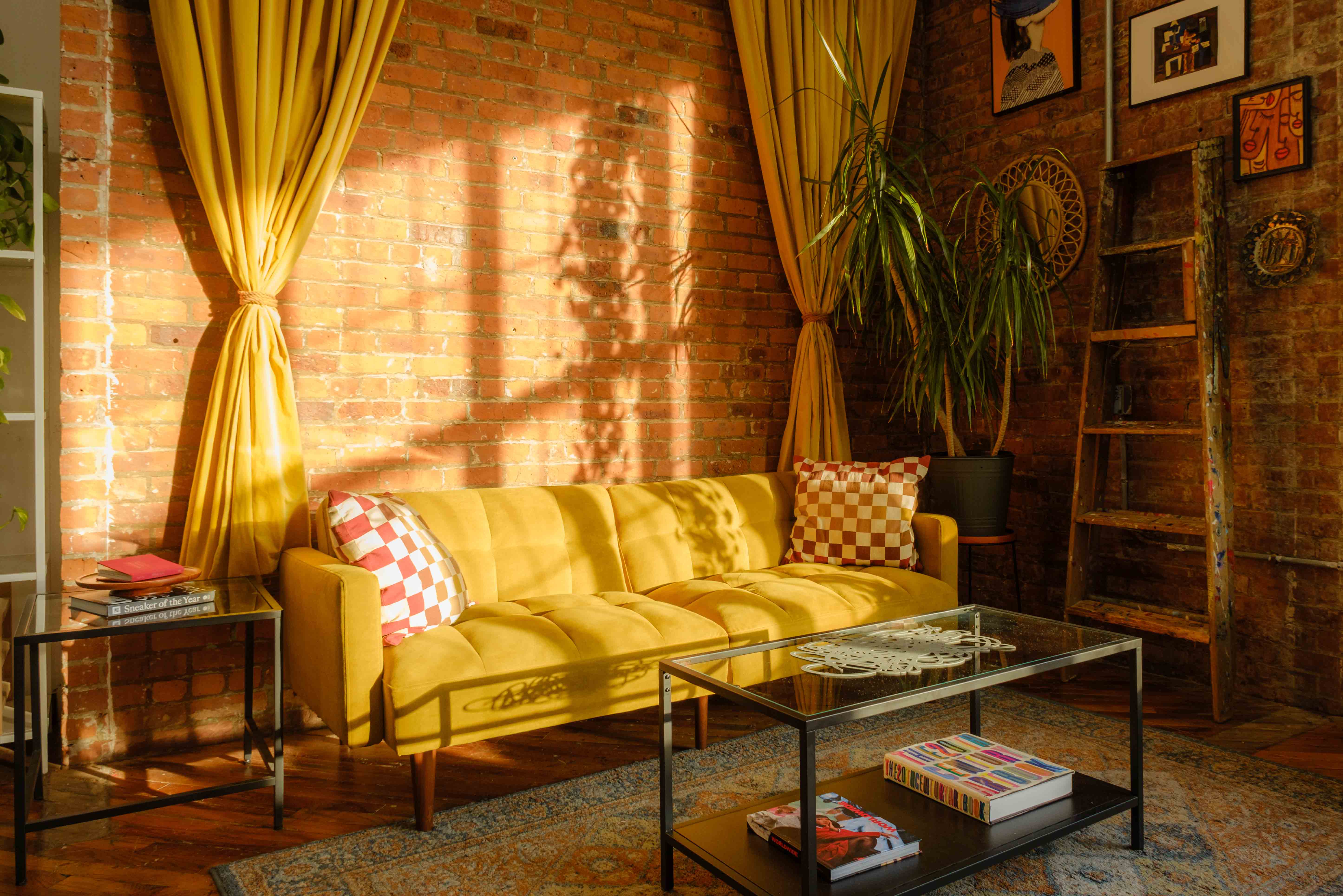 A cozy corner of a daylight photography studio with a yellow couch adorned with checkered pillows, set against an exposed brick wall. Bright yellow curtains and a potted plant add warmth and greenery, while a coffee table holds magazines, creating an inviting setting.