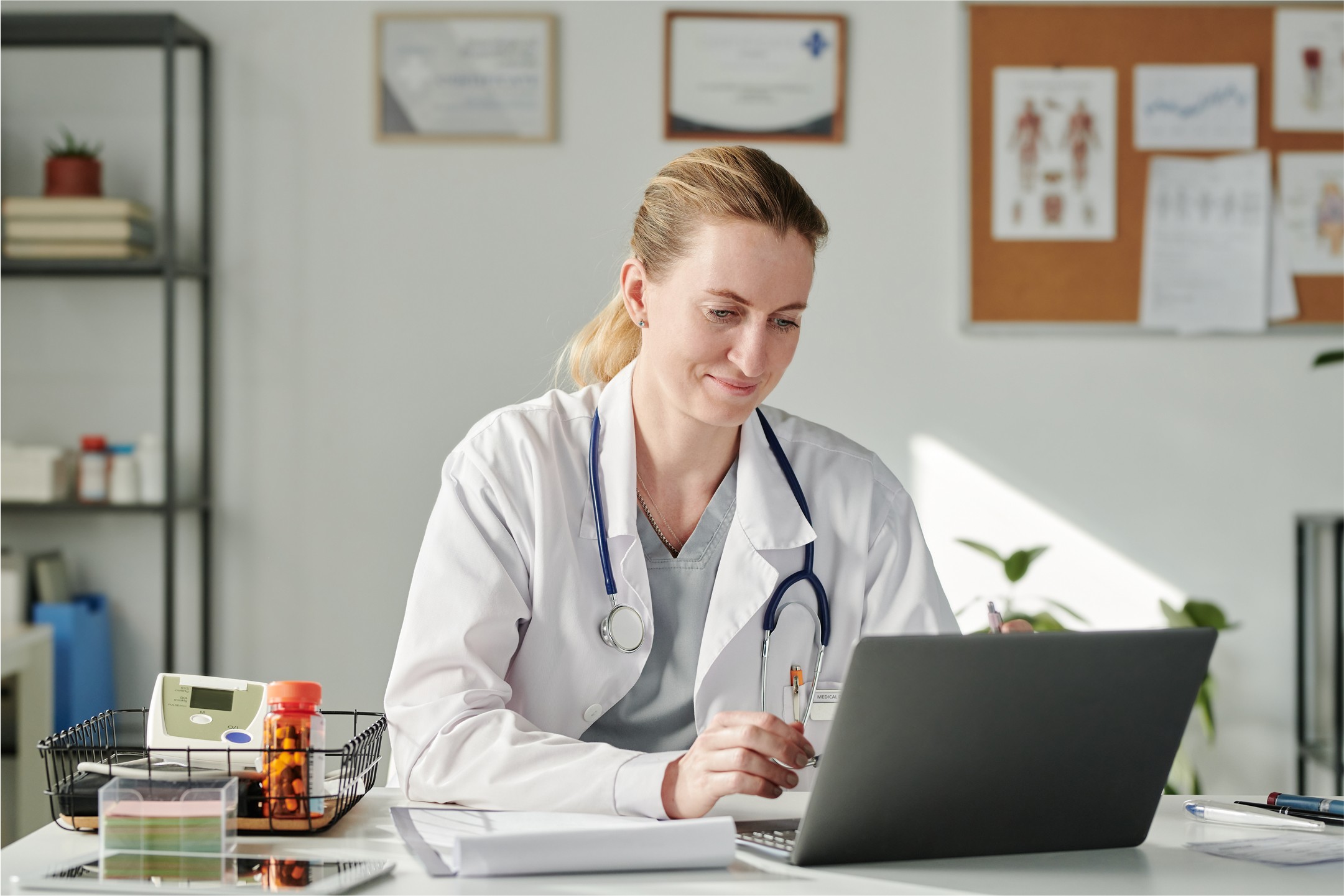 Female doctor using a laptop in her office, wearing medical scrubs.