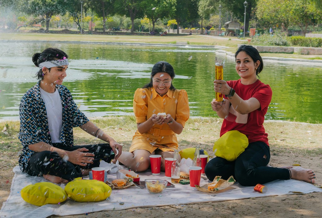 Three friends enjoying a festive picnic by the lake, laughing and playing with confetti poppers, surrounded by snacks and drinks on a blanket. The scene captures the joy and fun of outdoor gatherings and celebrations.