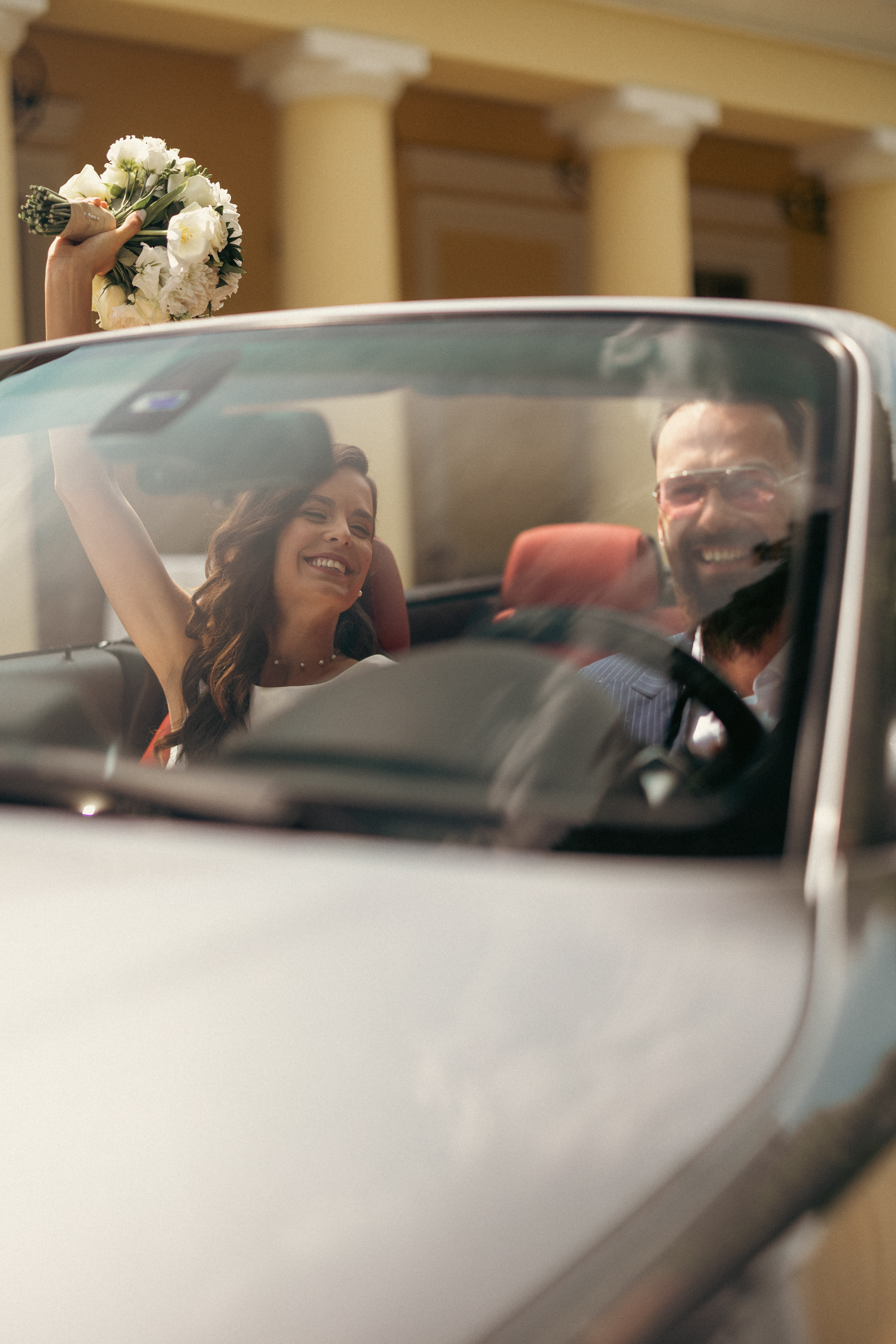Esküvői pár kabrió autóban, elegáns ruhában, spontán pillanatban.