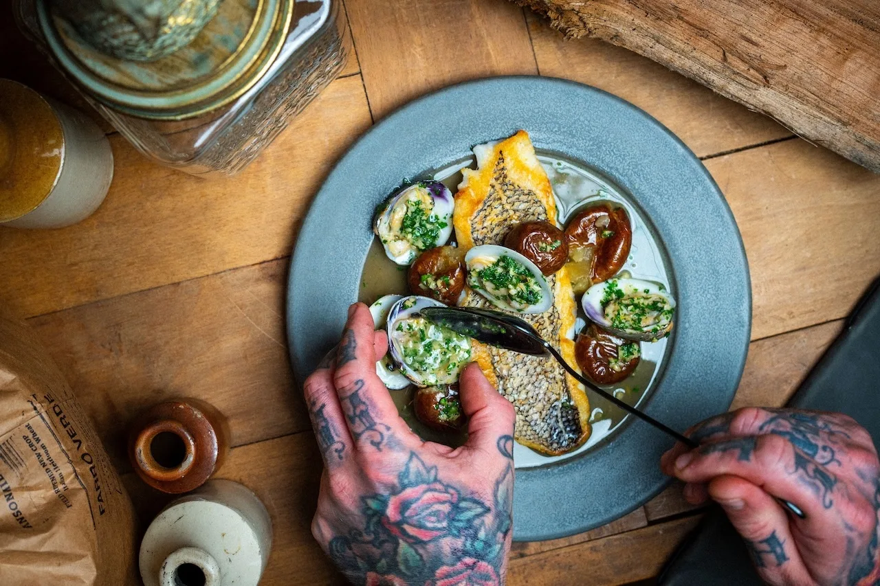 Black Briar promotional photograph of fish and clams on a blue plate, with the chef’s tattooed hands finishing the dish.