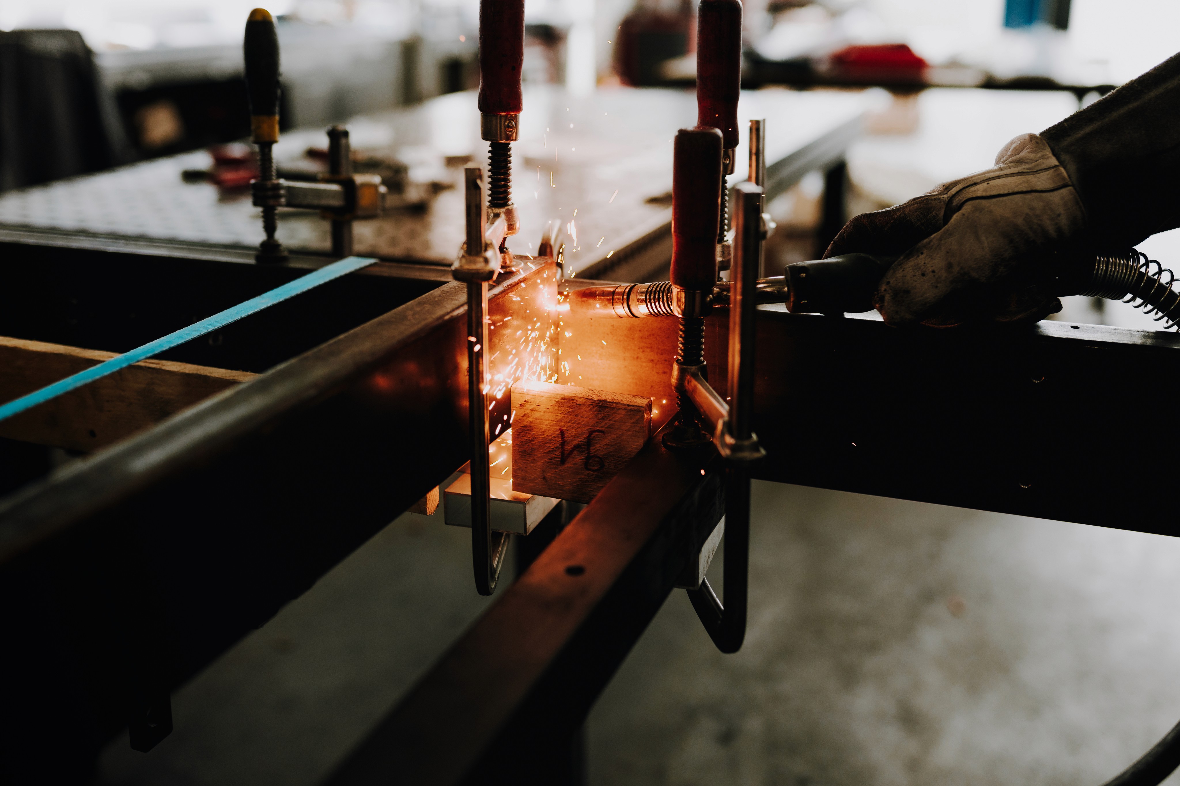 Close up of welding, showing sparks, clamps, a gloved hand and a metal structure.