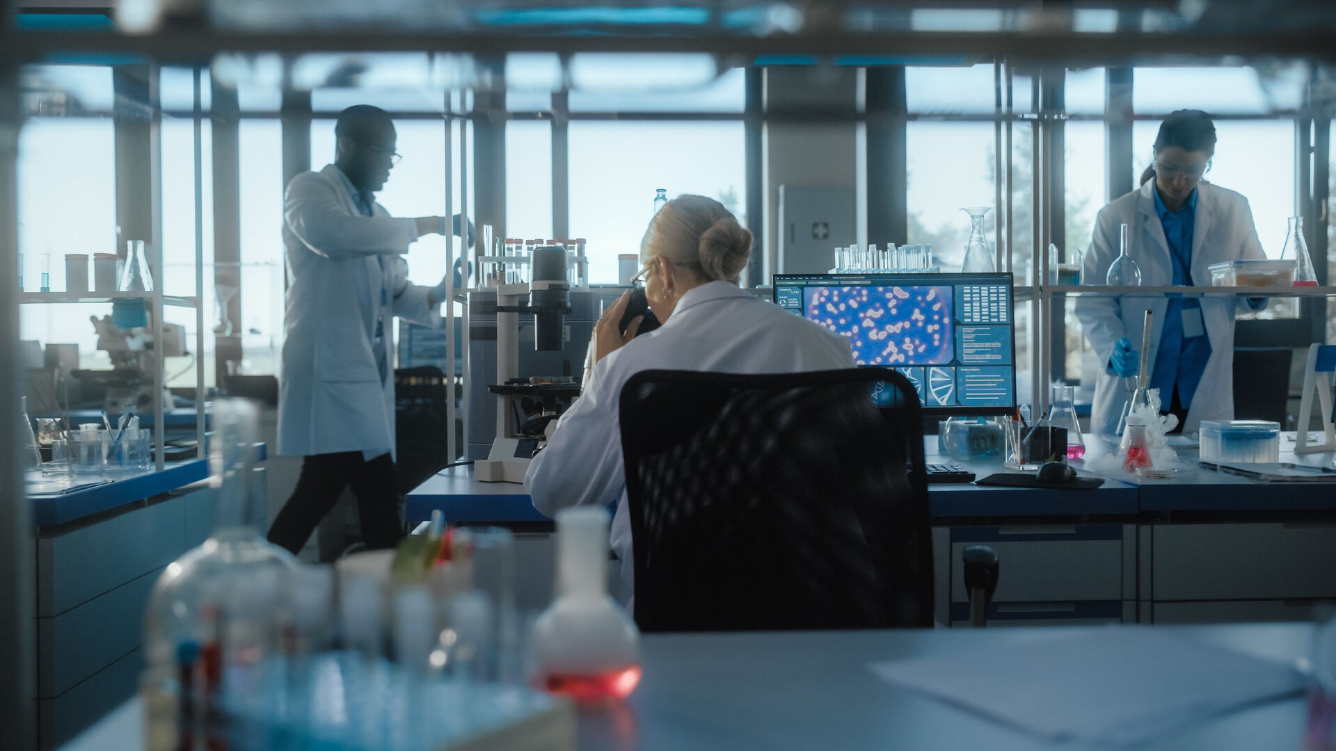A man in a white lab coat working at a computer, viewing a 3D model on the screen in a laboratory or office setting.