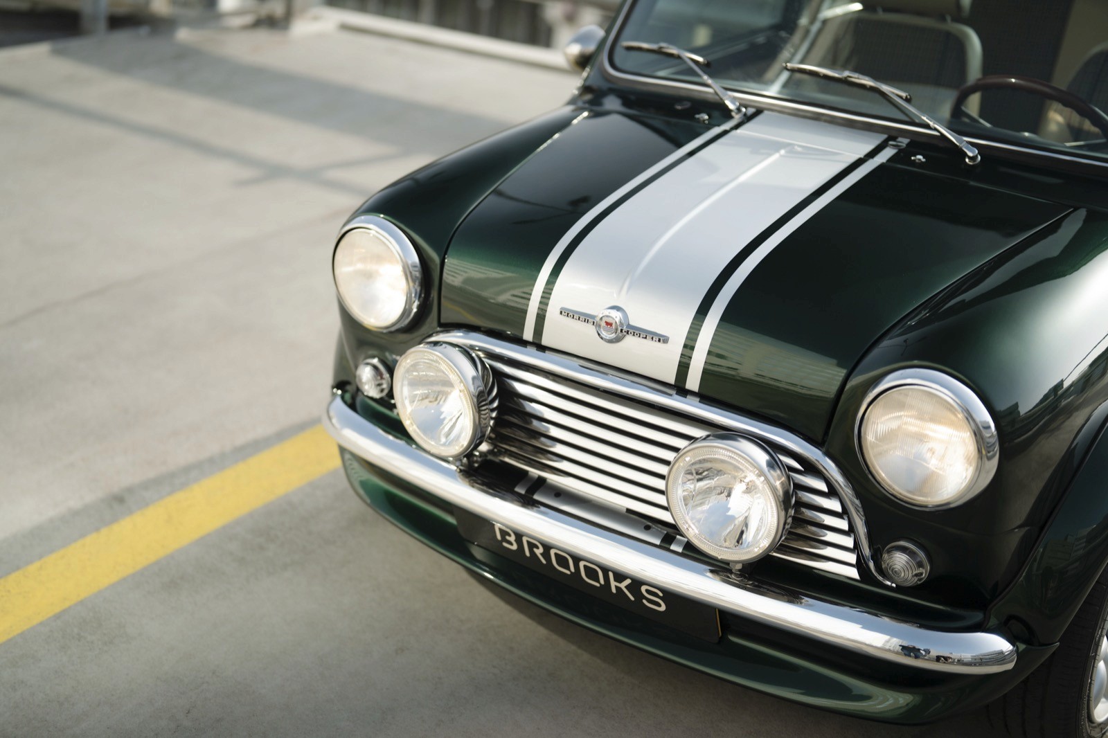 Close-up of a classic green car with white racing stripes and round headlights, parked on a concrete surface.