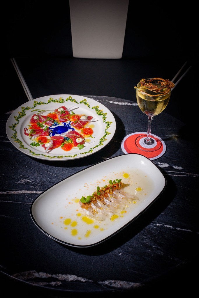 Two elegantly plated seafood dishes on a dark marble table with a cocktail beside them.