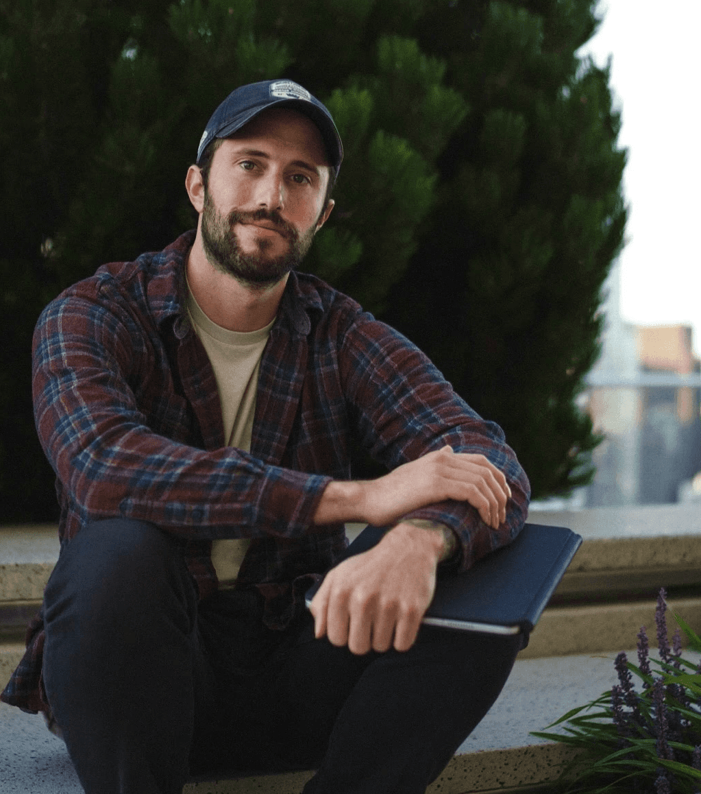 Person wearing a blue puffer jacket and red backpack standing in front of a light blue building on a city street.