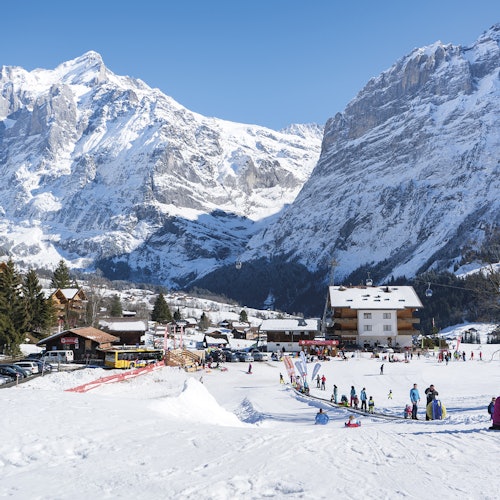 在白雪覆盖的滑雪胜地，人们正在滑雪和单板滑雪。周围是建筑物和高耸而崎岖的山脉，映衬着清澈的蓝天。