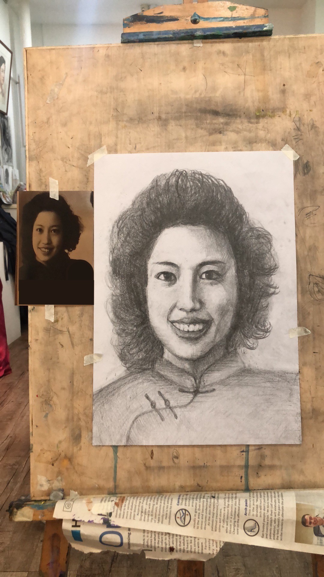 Pencil portrait of a smiling young woman with wavy hair, on an easel beside the photo it was drawn from