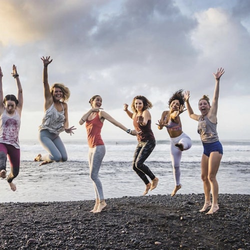 六位女性在岩石海灘上跳躍並快樂地微笑，背景是海洋和多雲的天空。