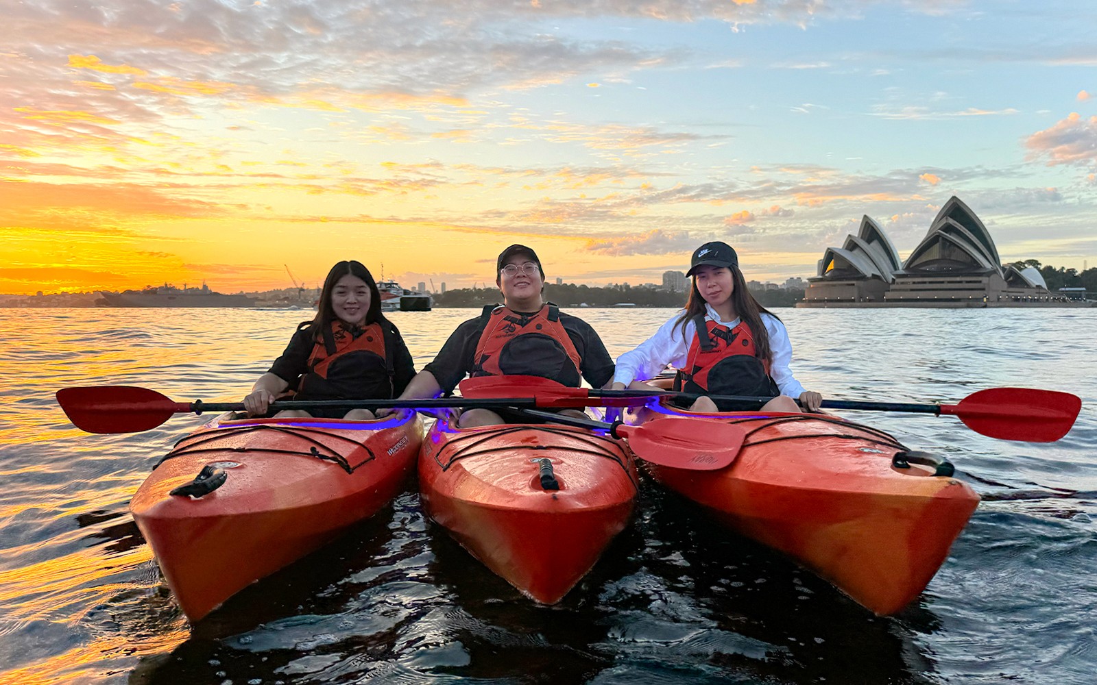 Group of people enjoying a sunrise paddle on Sydney Harbour with a breakfast spread on the kayak, with the iconic Sydney Opera House in the background