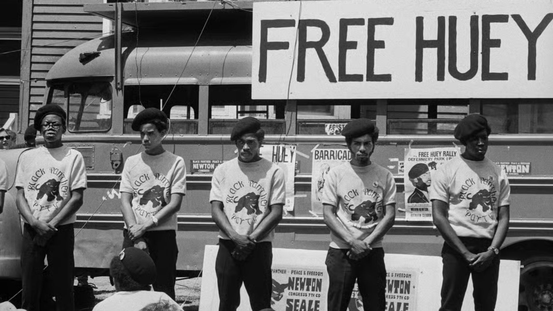 A black and white image of a 5 black panthers in front of a sign that says free huey on a bus all wearing black panther graphic tees.