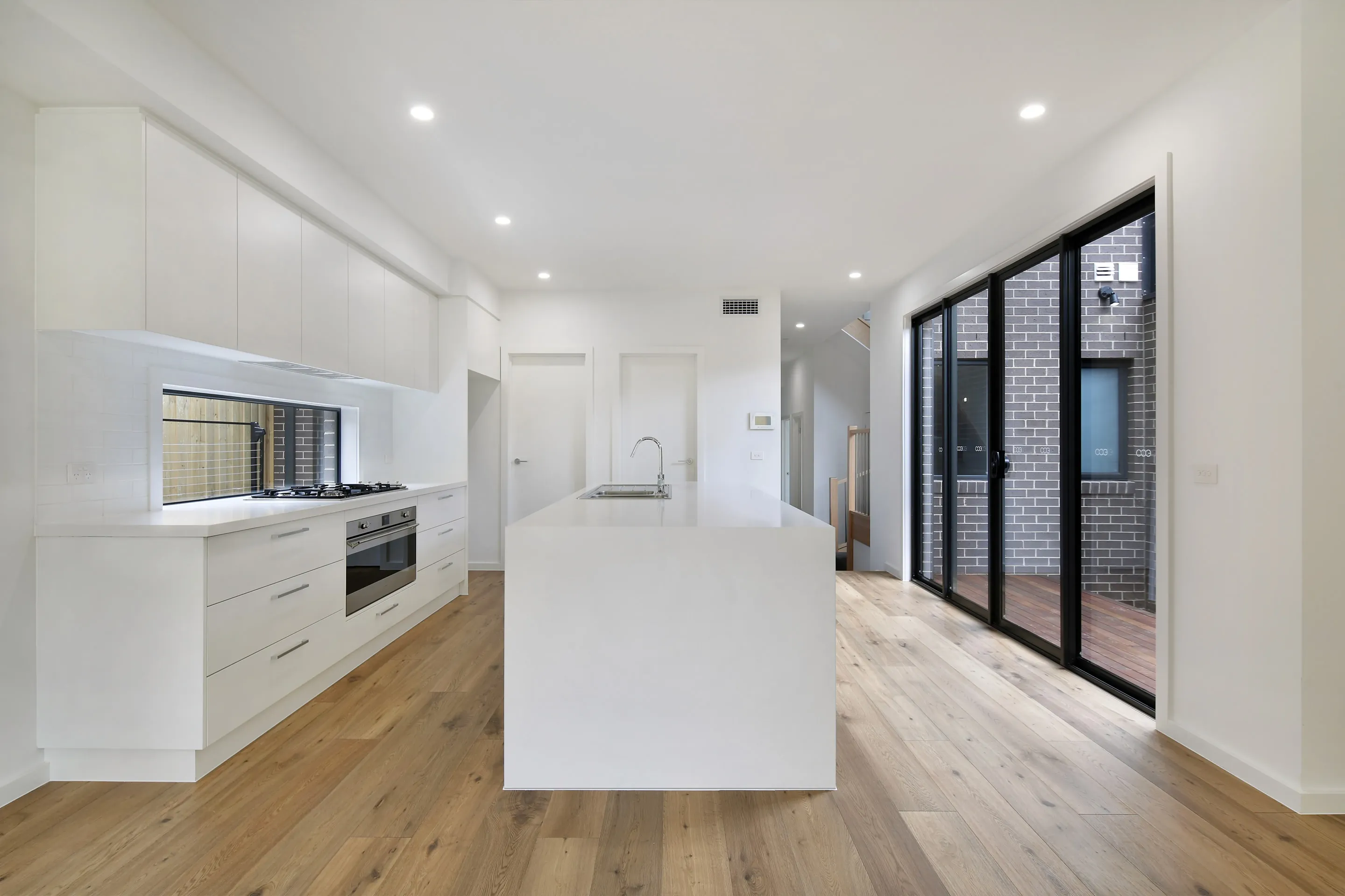 White galley kitchen with a waterfall island, overhead cabinetry, gas cooktop, integrated oven and chrome tap, beside black-framed sliding doors to a brick courtyard and a stair beyond