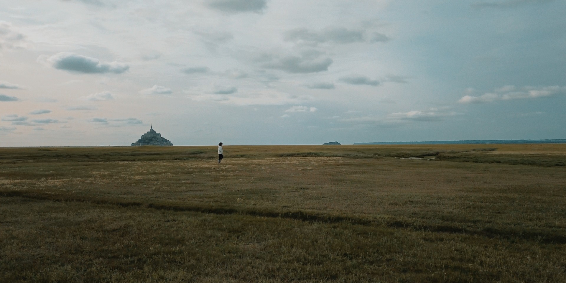 Vue panoramique d’un promeneur solitaire devant le Mont-Saint-Michel sous un ciel nuageux.