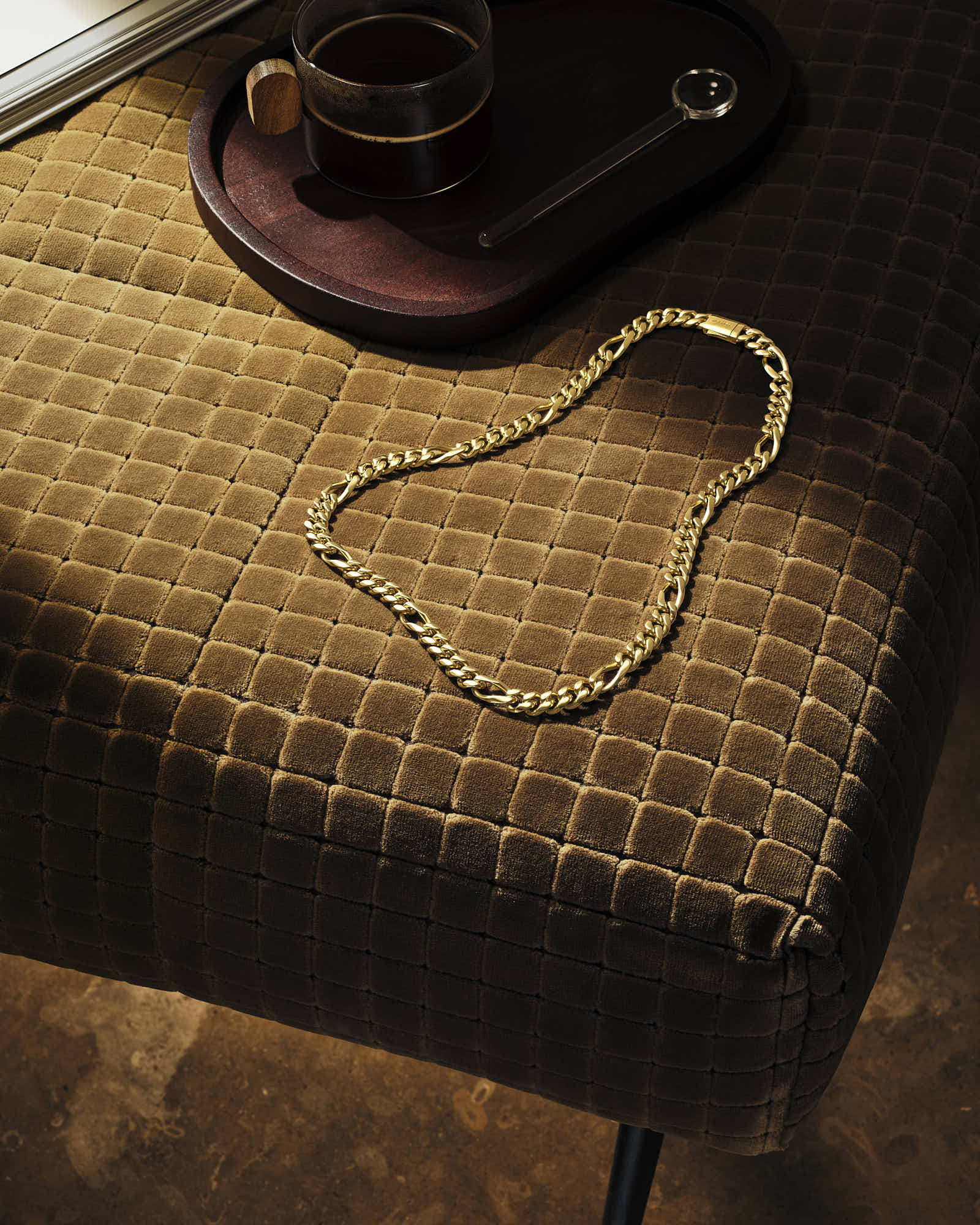 Gold chain necklace on brown textured sofa with wooden tray and coffee cup.