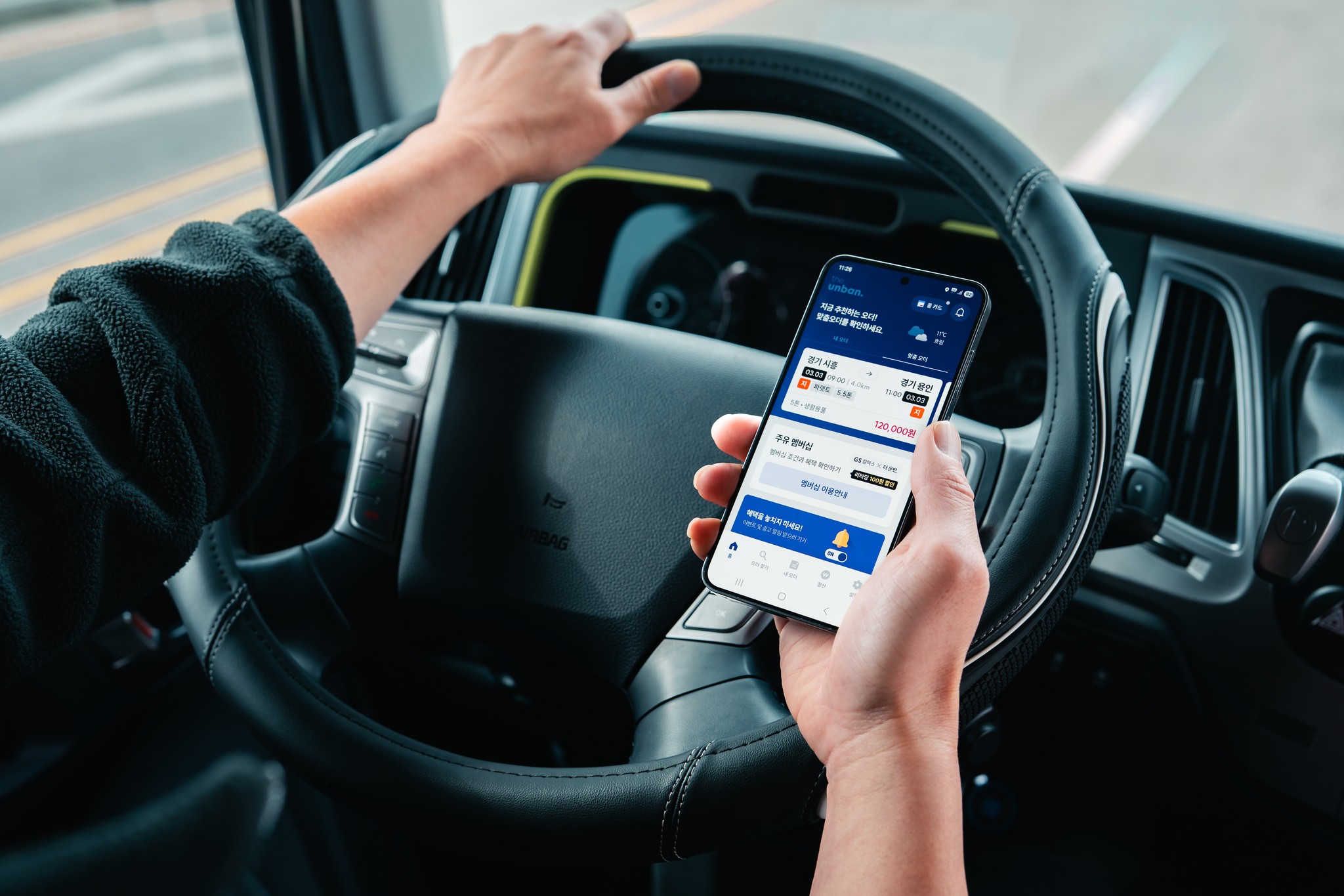 CJ대한통운 더 운반 차주 앱을 소개하는 이미지로, 운전석에 앉은 차주가 운송 오더와 운임 정보가 표시된 더 운반 앱 화면을 들고 있습니다. 배차부터 운임 정산까지 앱 하나로 처리할 수 있다는 메시지와 함께 앱 가입 버튼이 안내되어 있습니다.