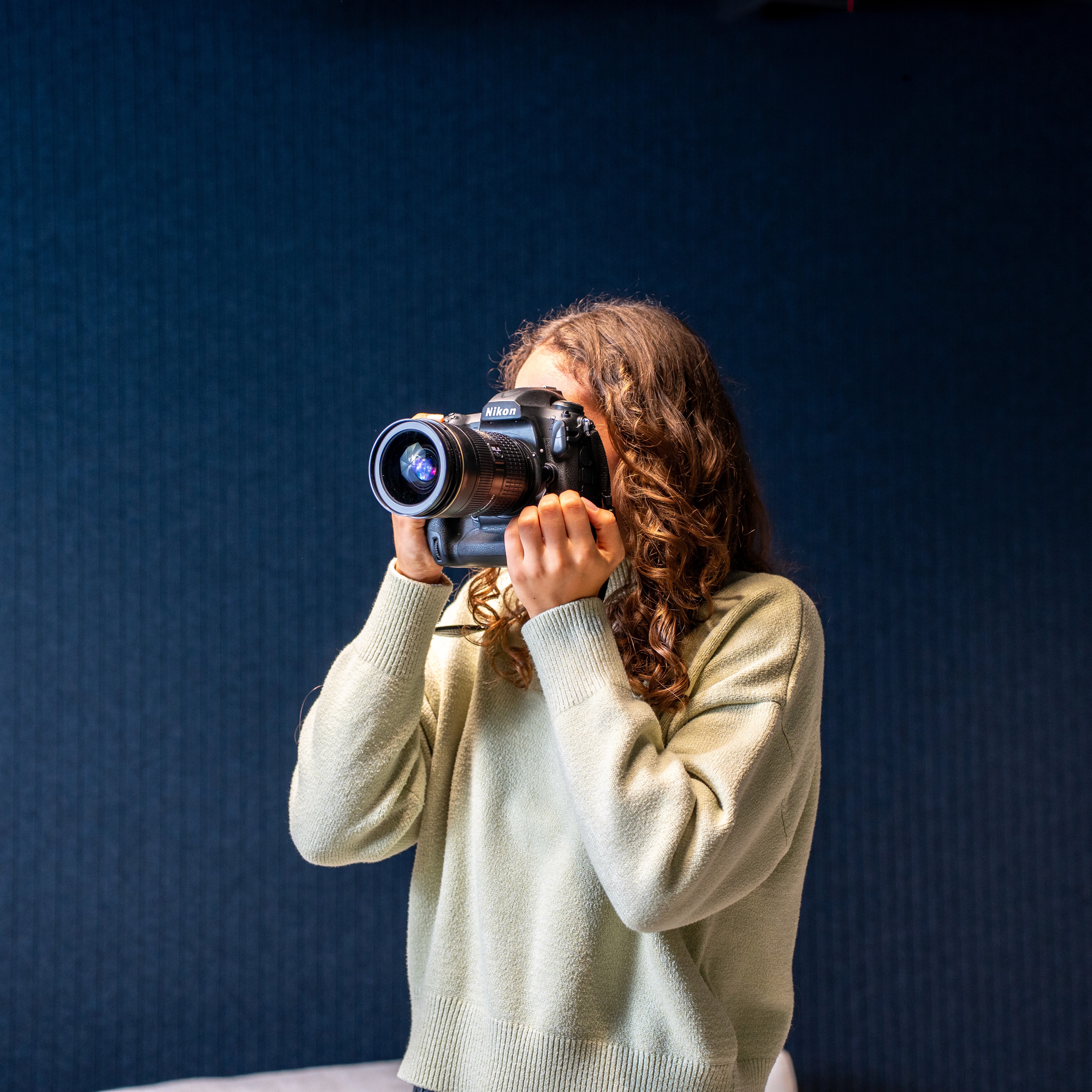 Smiling young woman with long hair standing against a dark green background, holding a finger to her chin.