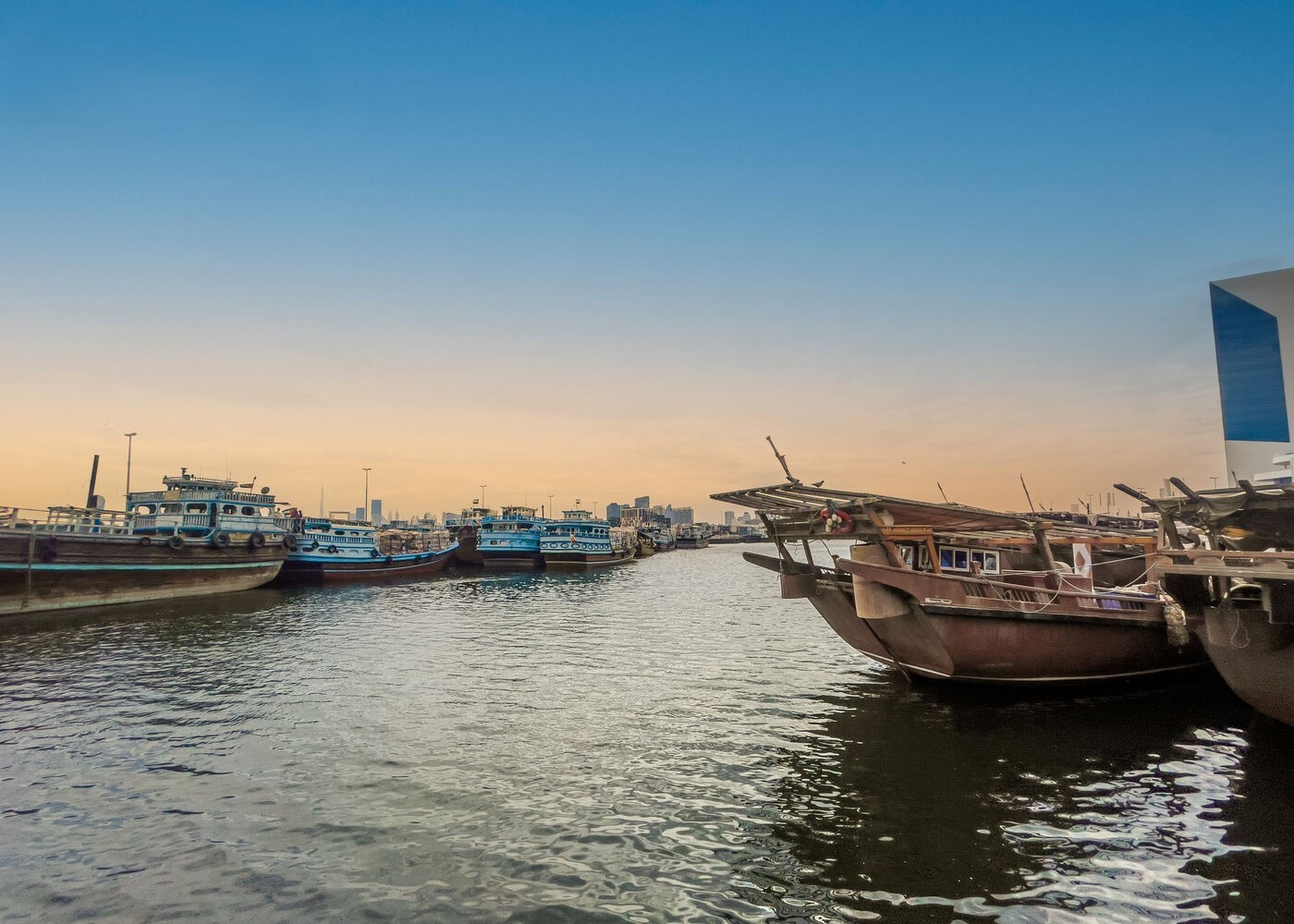 Sunset over Deira harbor with moored boats, calm water, and cityscape blending traditional vessels with urban skyline.