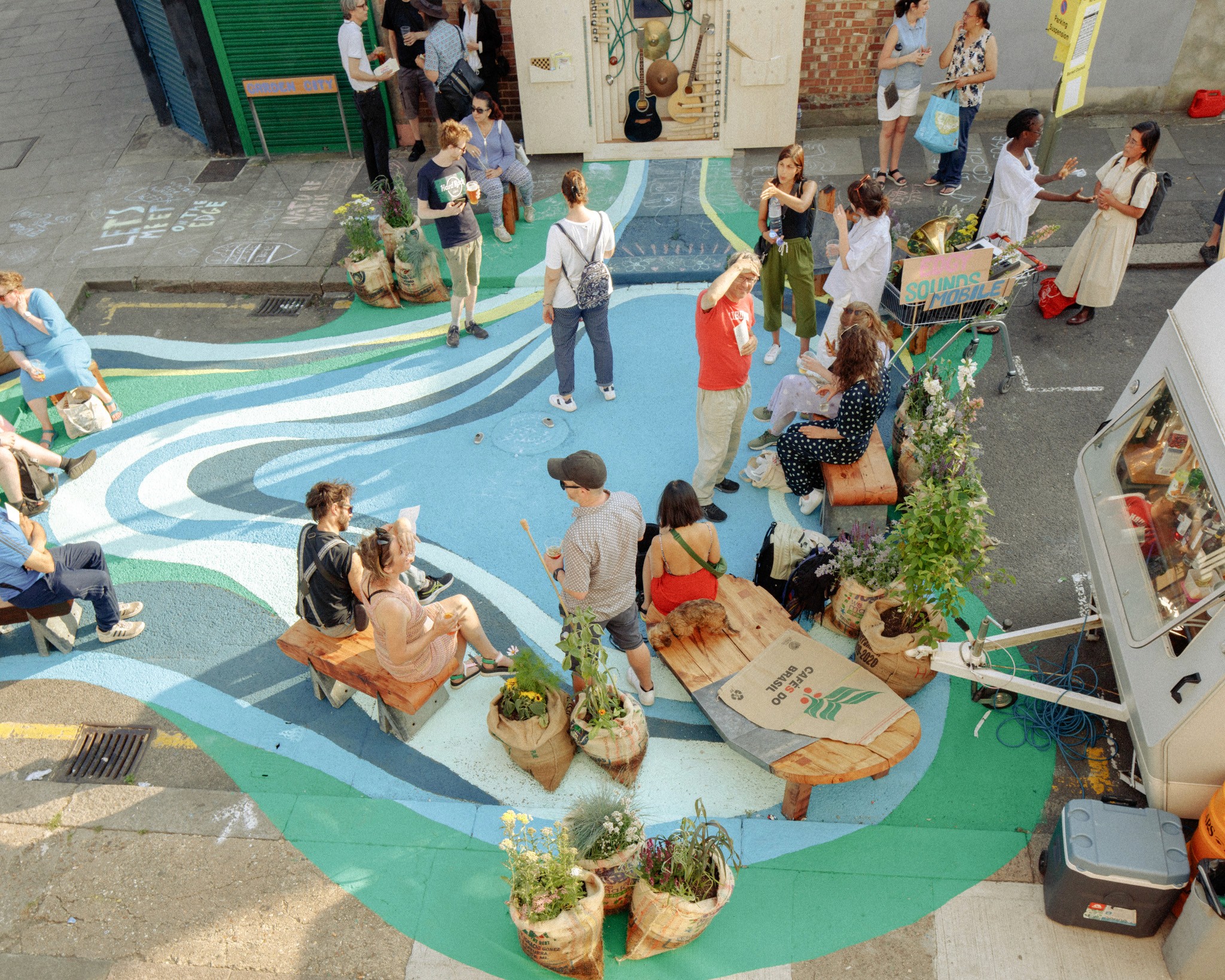 Overhead view of a community street gathering with people both sitting and standing around a colourful ground mural, as well as plants and instruments