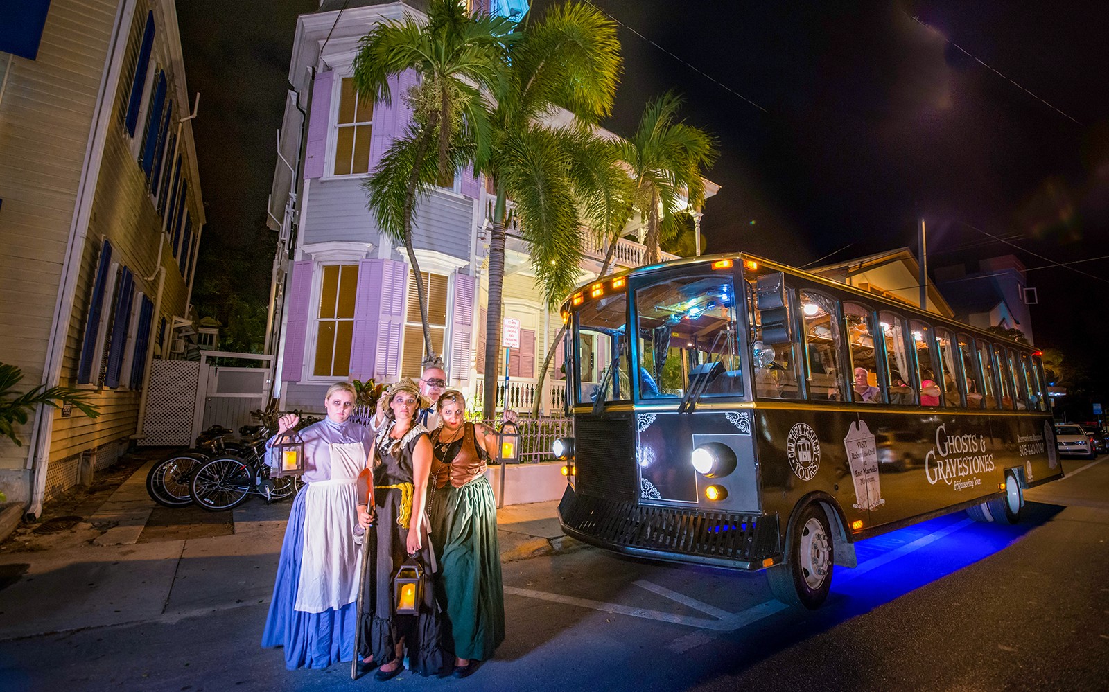 Tour group in historical costumes with lanterns beside Boston Ghosts & Gravestones trolley at night.
