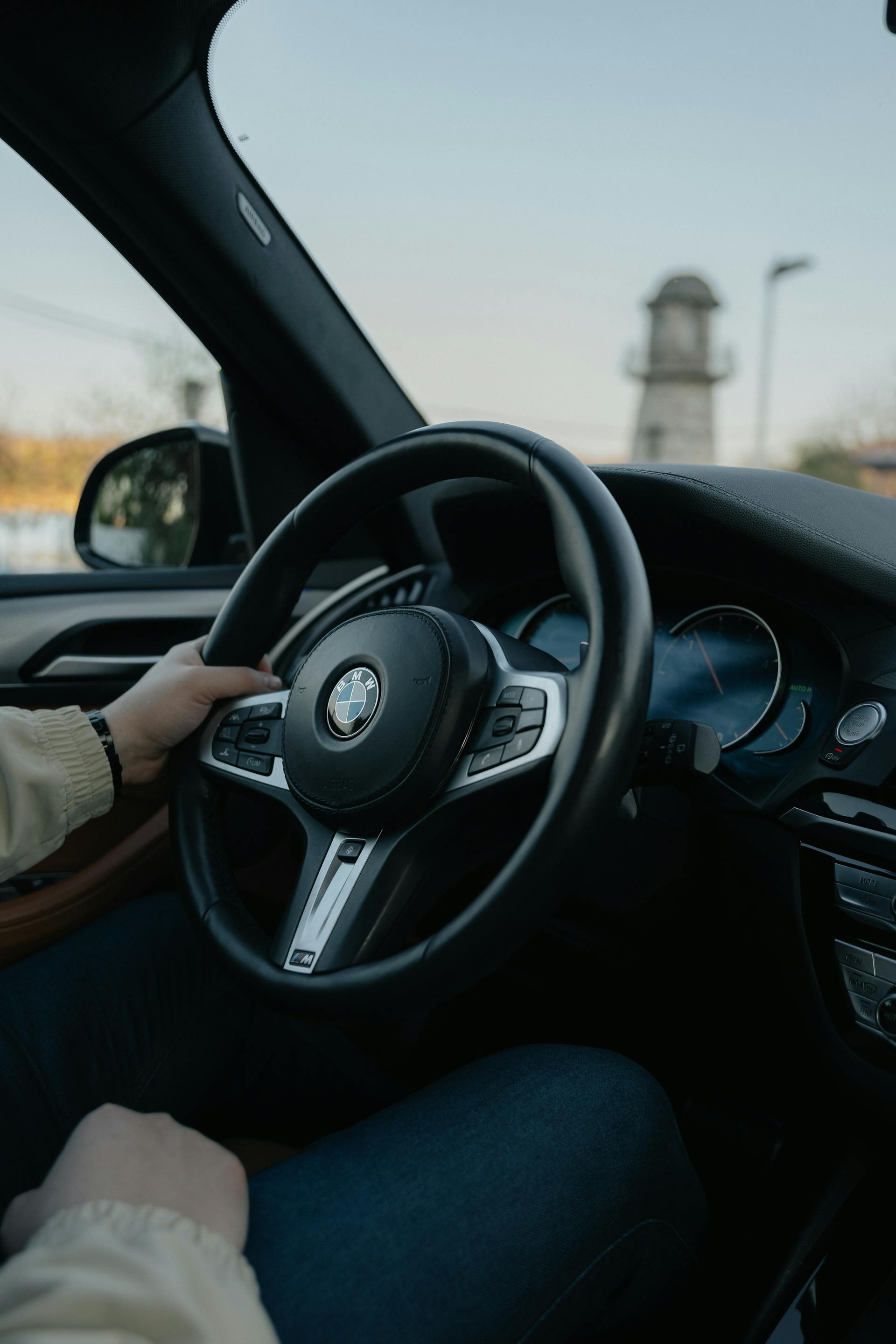  A close-up shot of a person's hands and lap inside a modern car, likely a BMW (indicated by the logo on the steering wheel). The person is wearing a light-colored sweater and dark jeans. The driver's left hand is resting on the steering wheel. Through the windshield, a stone tower or lighthouse is visible in the background against a light sky.