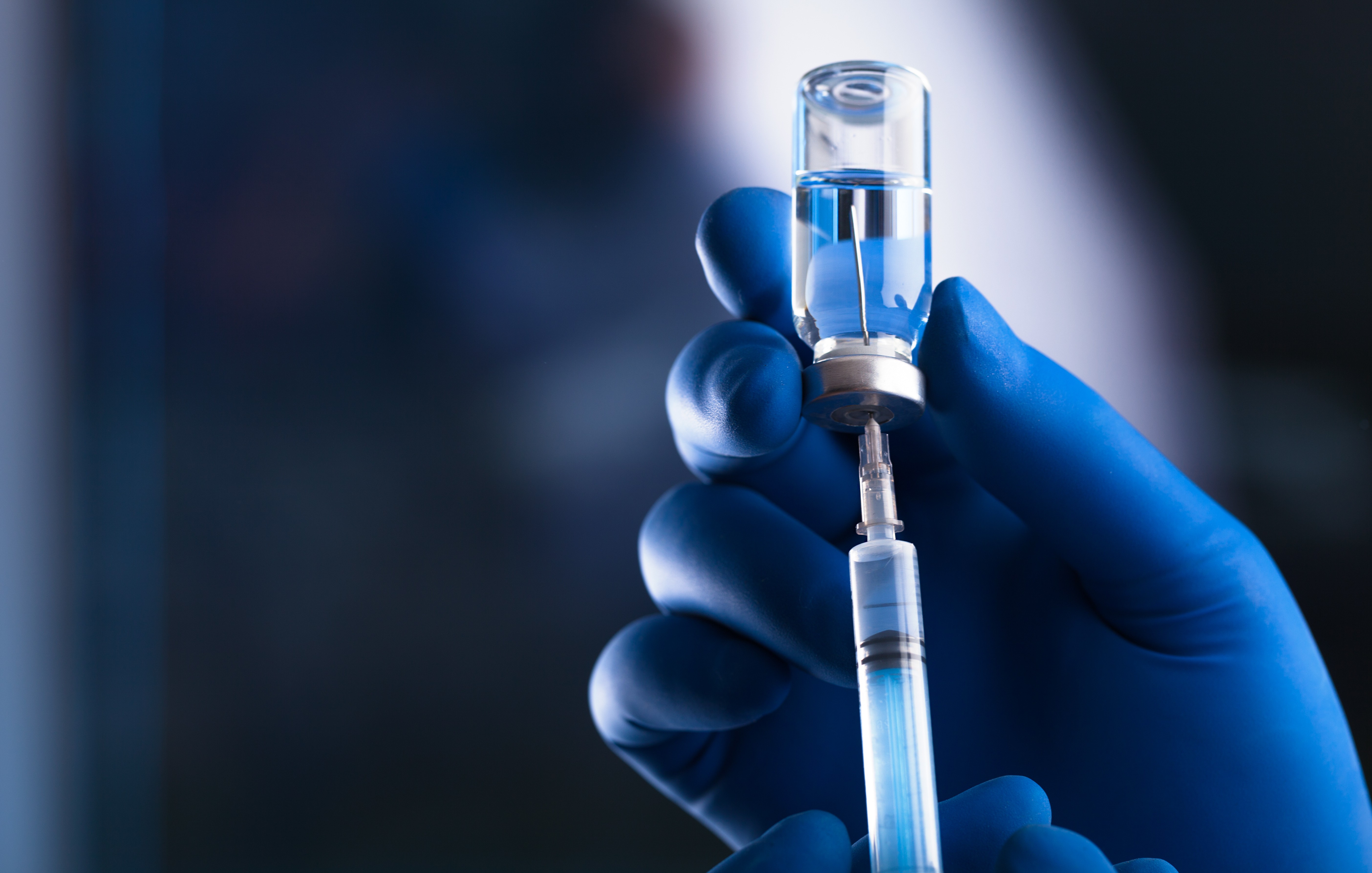 A hand wearing blue gloves holding a vial that is upside-down with a syringe and a needle inserted into it to extract the clear liquid inside. 