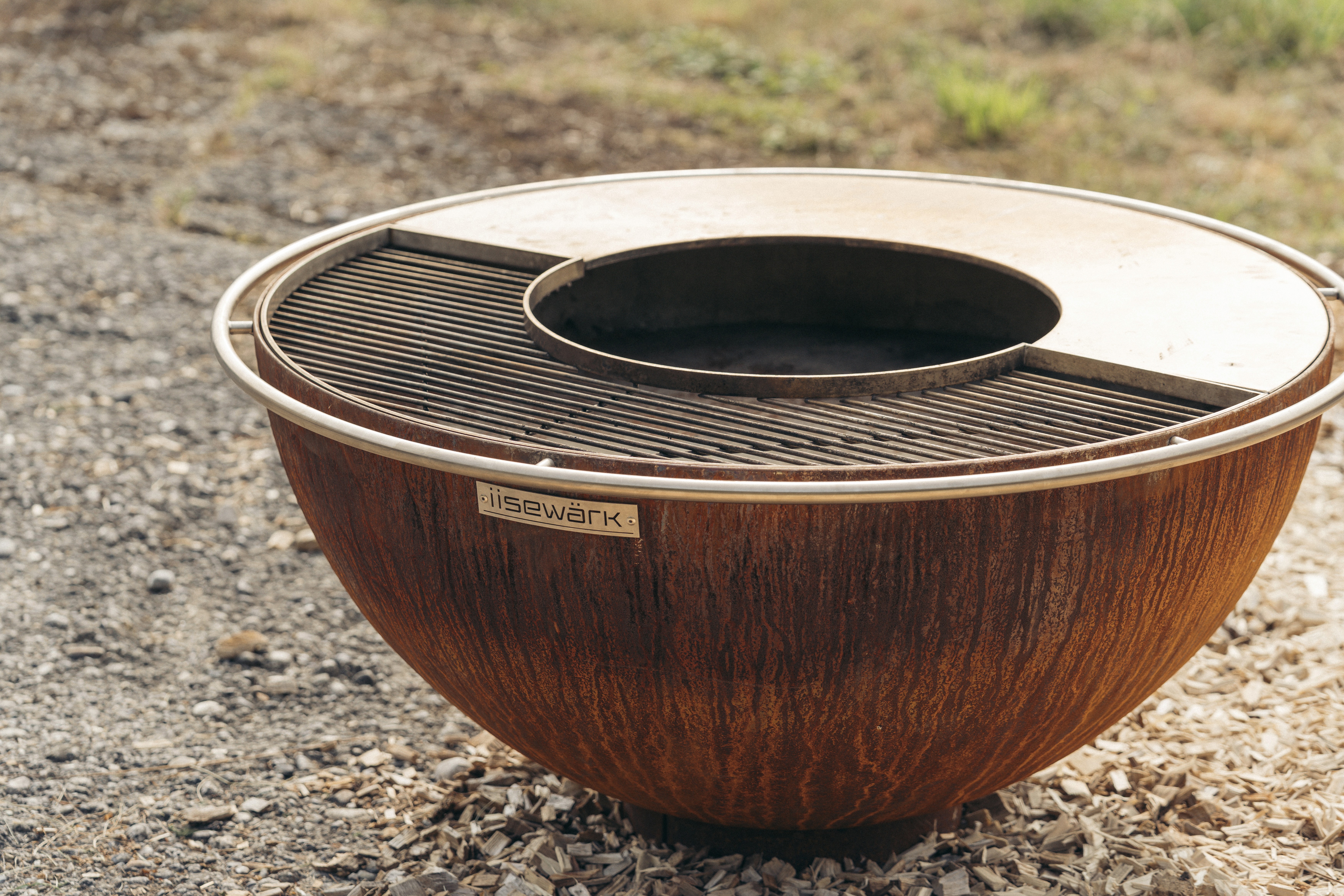 Eine große, runde, rostfarbene Schale steht auf einem Steinsockel, umgeben von einer natürlichen Landschaft aus Grünpflanzen.