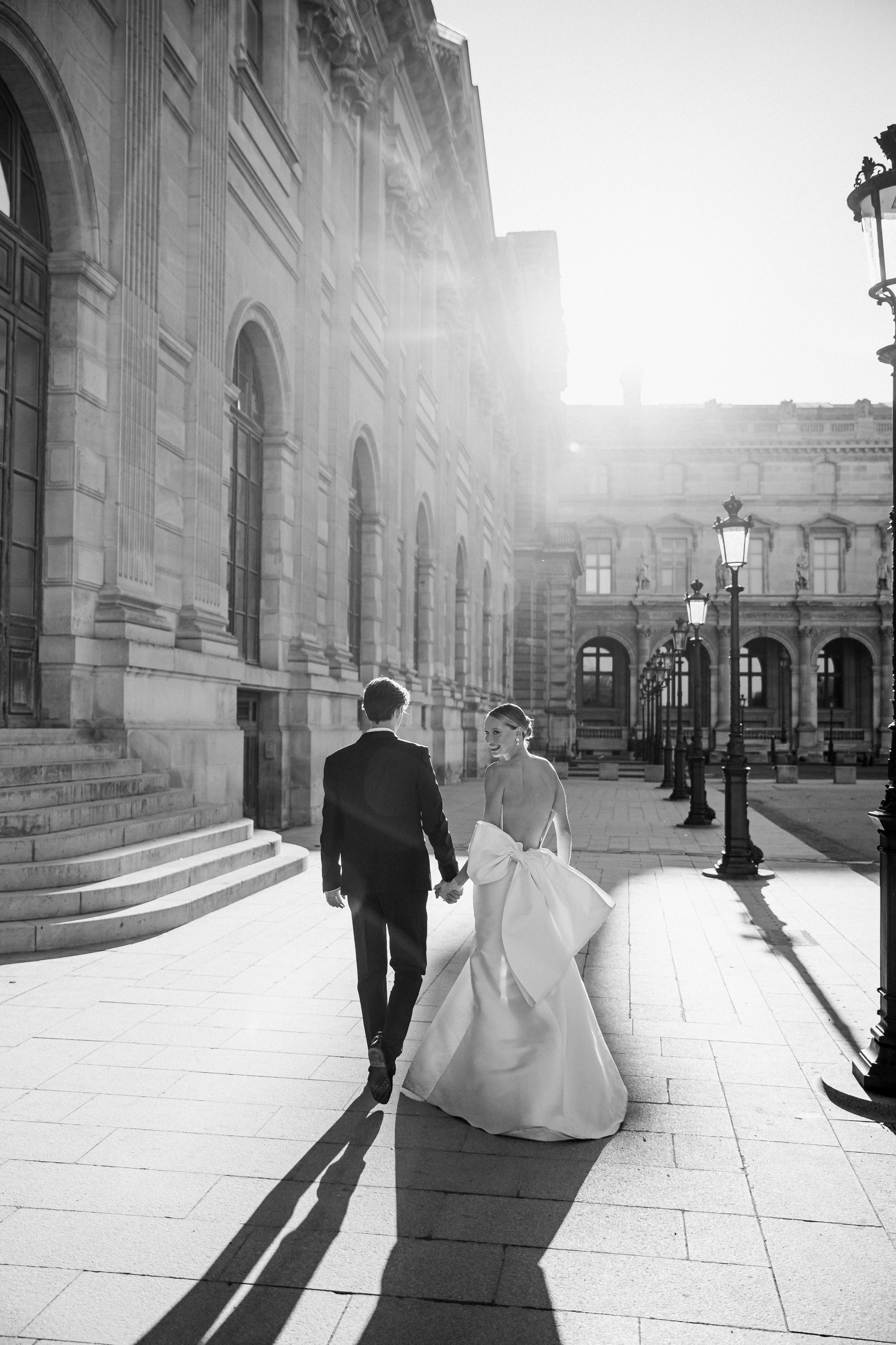 A groom in a dark suit carries a bride in a white dress across a sunny, grassy field. The couple shares a joyful, intimate moment with trees in the background.