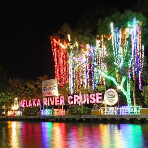 Colorful lights illuminate trees and a "Melaka River Cruise" sign at night, reflecting off the water below.