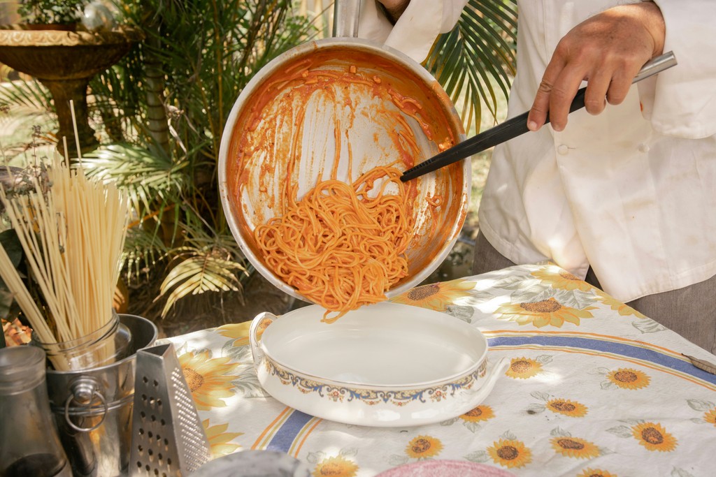 A chef in a white coat serves freshly cooked spaghetti in a rich tomato sauce onto a decorative plate on a floral tablecloth, with uncooked pasta and a cheese grater nearby, set in an outdoor, sunny garden setting.