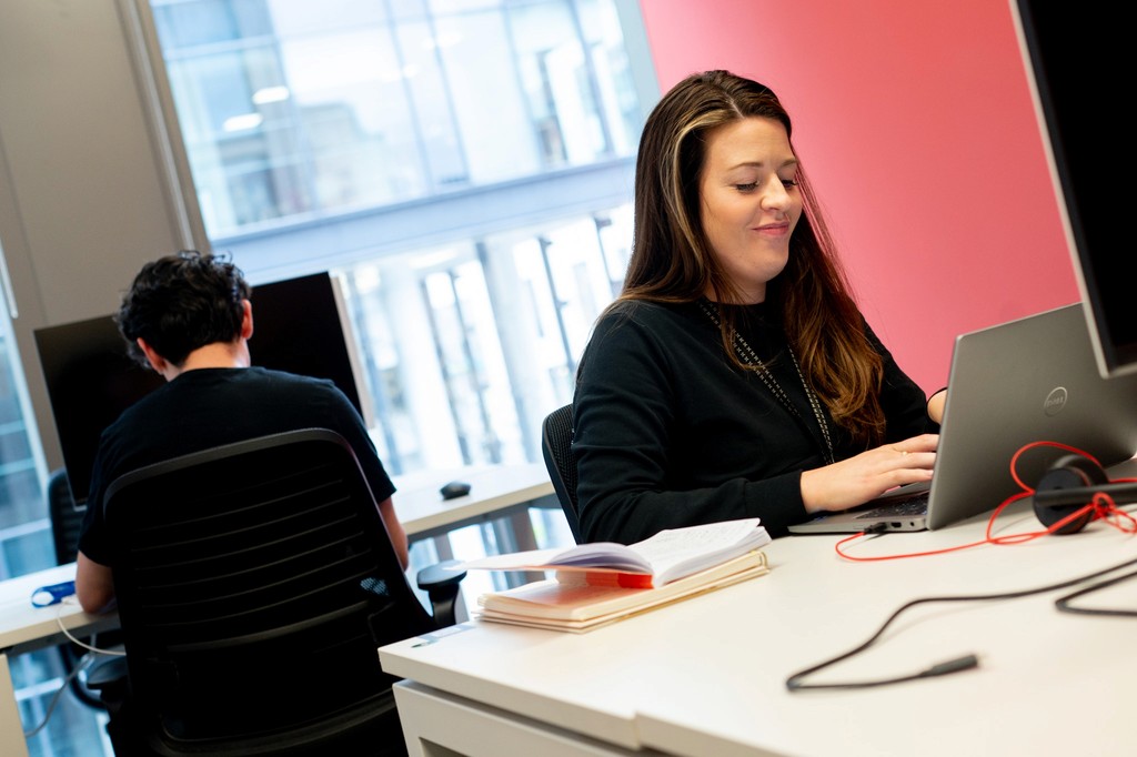 Woman working on laptop at desk, smiling. Another person at a desk in the background, working on a computer.
