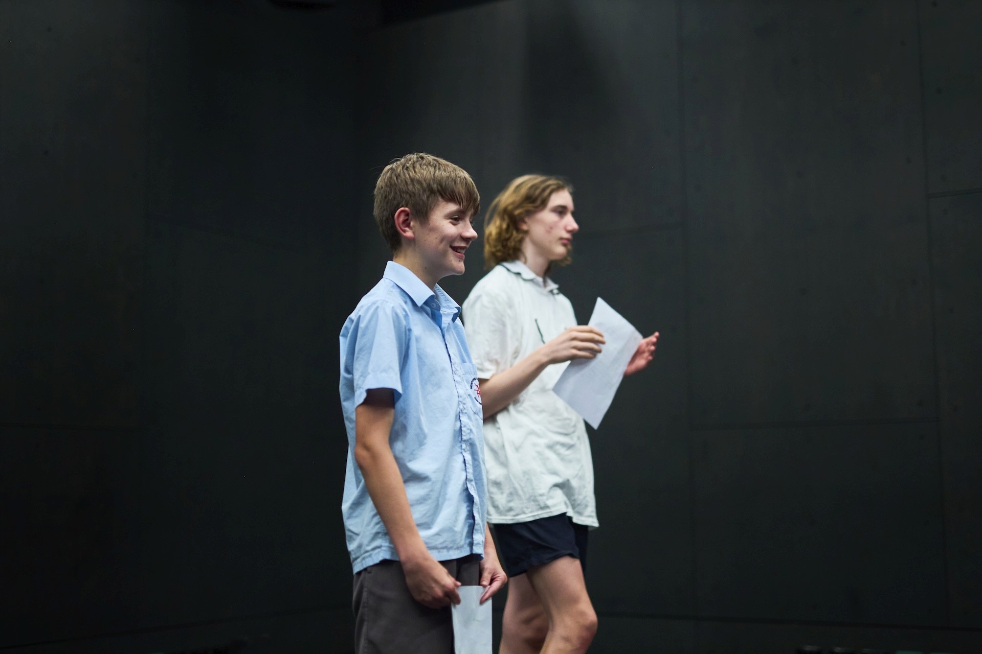 Two teenagers stand in a dark-walled studio holding scripts, preparing to read during a rehearsal.