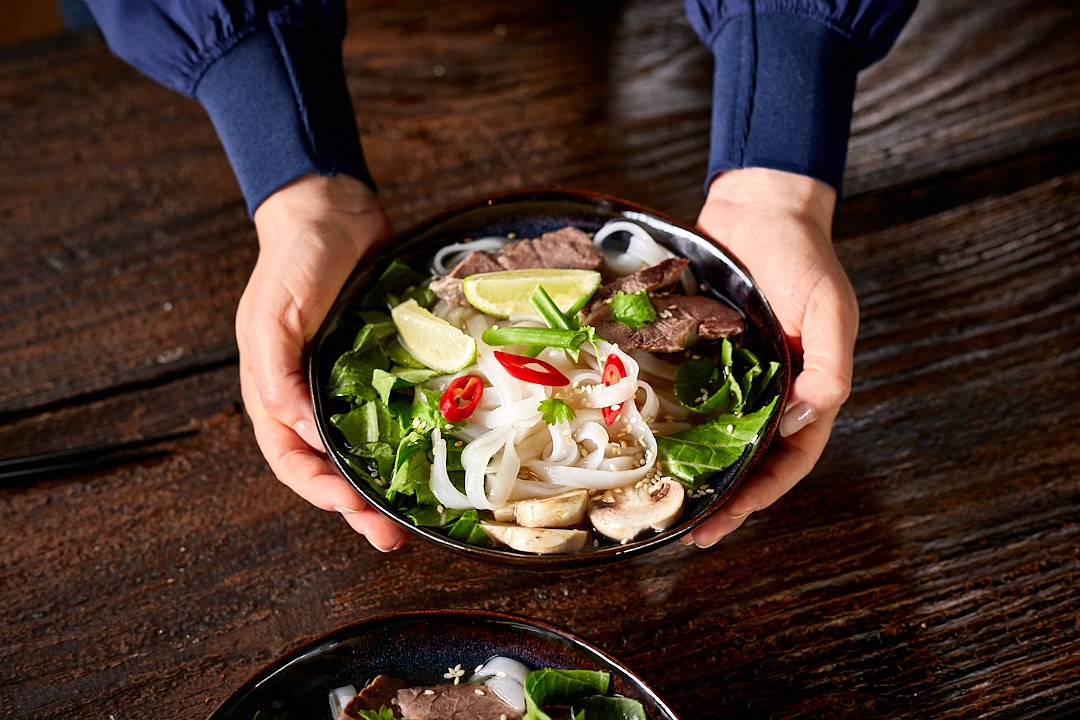 A person's hands serve a bowl of Vietnamese pho, featuring rice noodles, sliced beef, fresh herbs, lime wedges, sliced red chilies, and mushrooms, on a rustic wooden table.