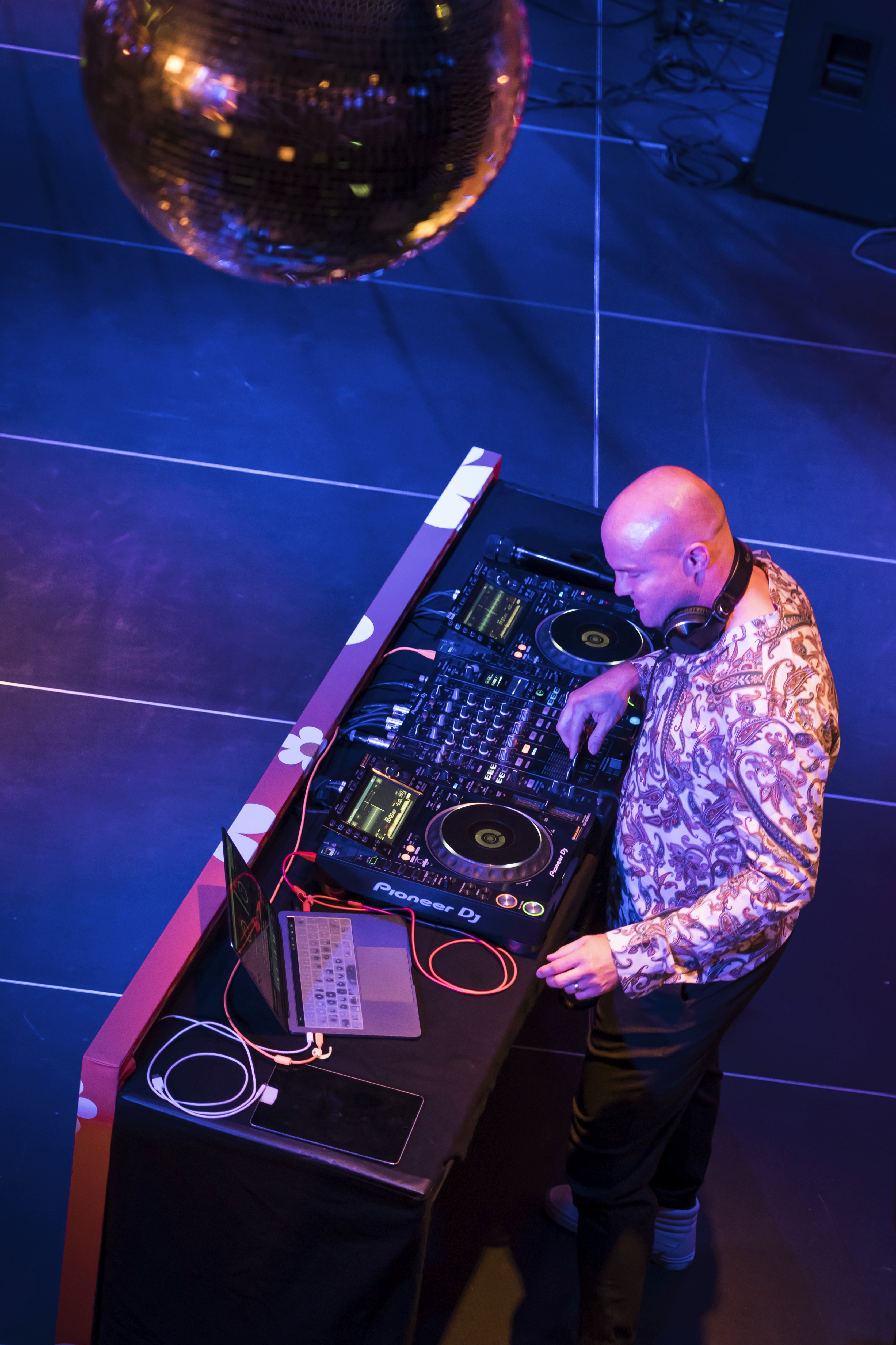 Das Foto zeigt aus der Vogelperspektive einen DJ bei der Arbeit an seinem Pult. Der glatzköpfige Mann trägt ein helles, auffällig gemustertes Hemd (Paisley-Muster) und hat schwarze Kopfhörer um den Hals gelegt. Er bedient konzentriert ein großes, professionelles DJ-Mischpult (der Marke Pioneer), neben dem auf dem schwarzen Tisch auch ein Laptop und ein Tablet liegen. Die Szenerie wird von stimmungsvollem, intensivem blauem und pinkem Scheinwerferlicht beleuchtet. Im oberen linken Bildbereich ragt eine große, schimmernde Diskokugel ins Bild.
