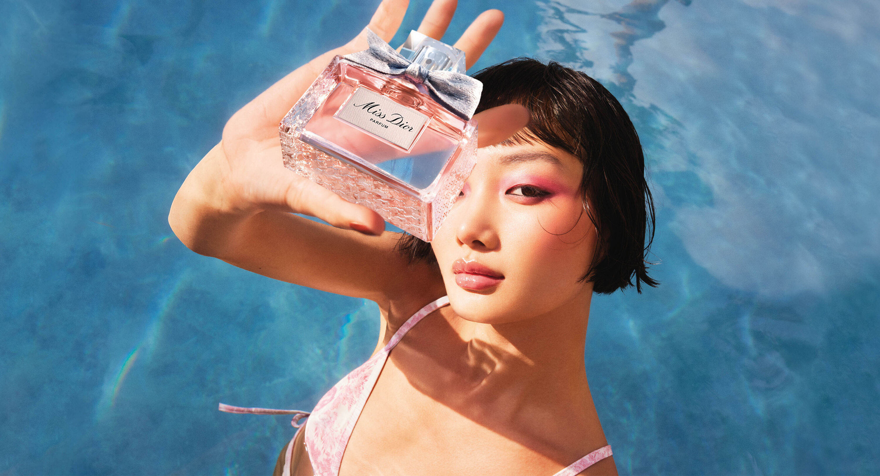 Model holding up a Miss Dior perfume bottle in front of her face, with bold pink eye makeup and a pool in the background for 'Dior Beauty'.
