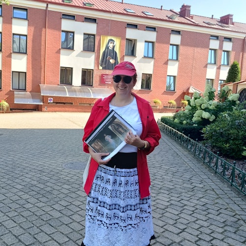 Женщина в красной кепке и рубашке держит папку на улице, на фоне кирпичного здания и религиозной картины.