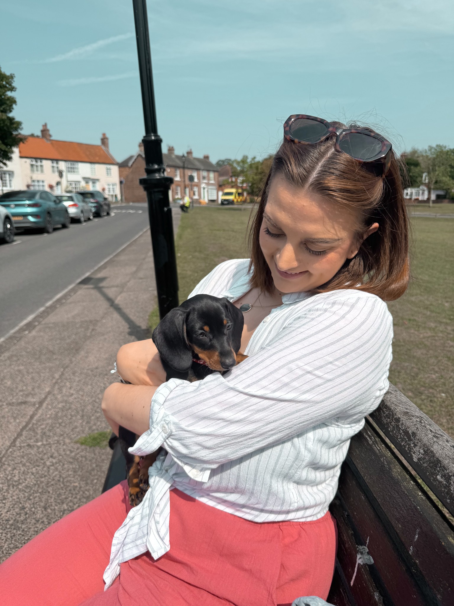 Card image of the business owner (Female late-20s, long brown bob haircut, wearing pinky/orange trousers and a white shirt) sat on a bench cuddling a black and tan Dachshund puppy