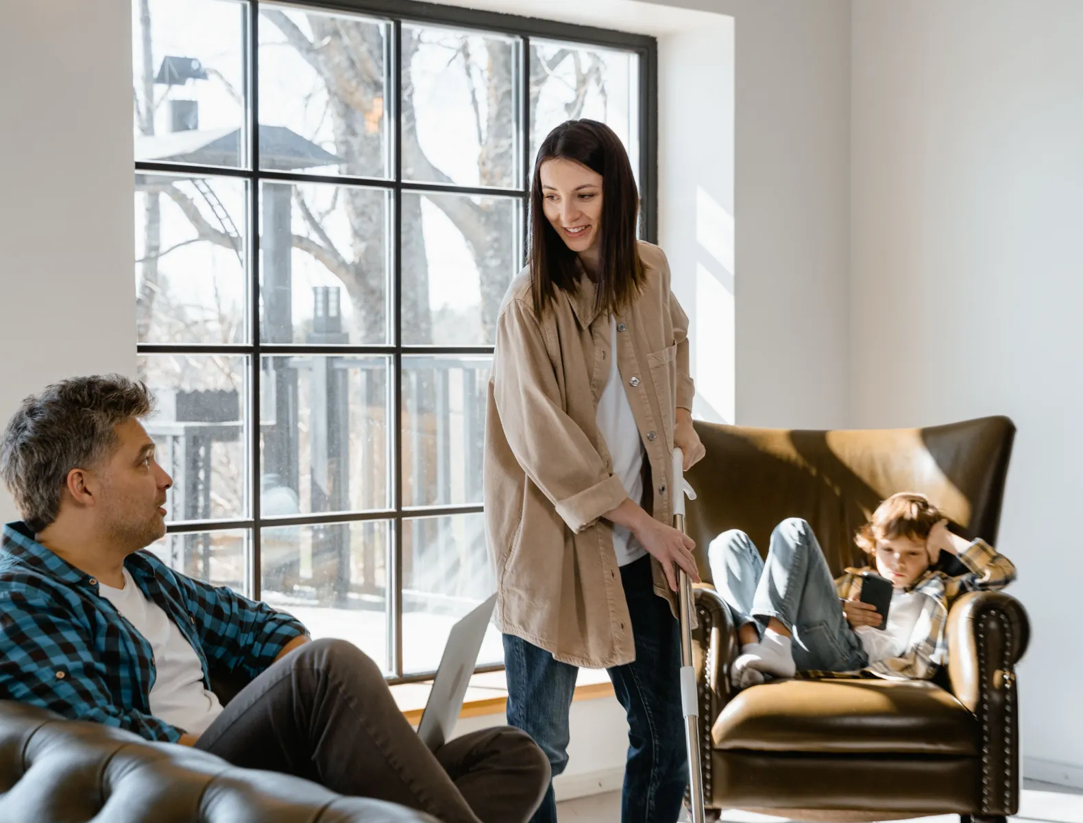 A family in a naturally lit living room. The father is sitting on a couch speaking to the mother who is vacuuming. Their child is sitting in an armchair in the background on a phone.