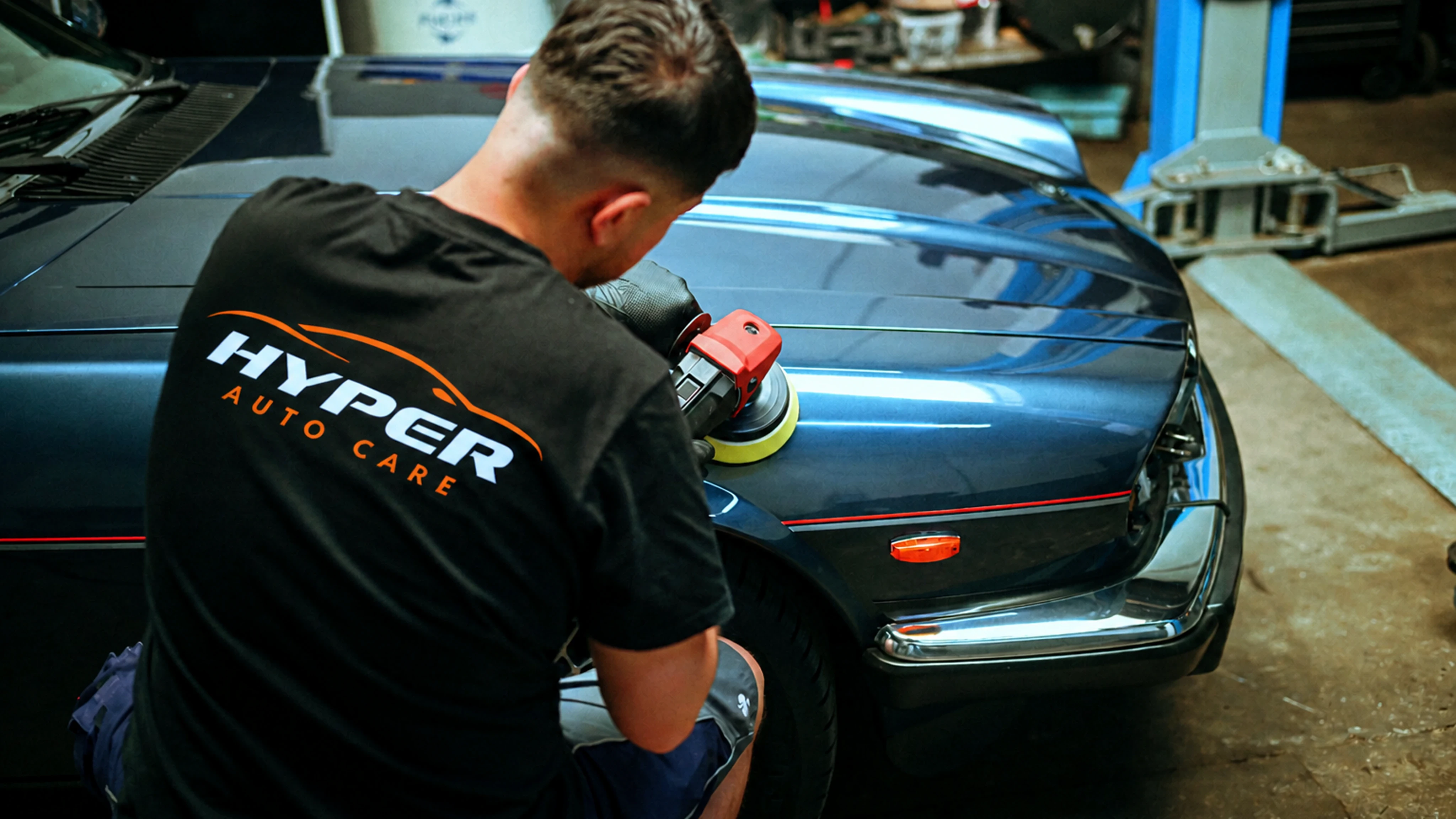 HyperAuto Care technician polishing a dark vehicle hood inside a professional auto detailing workshop