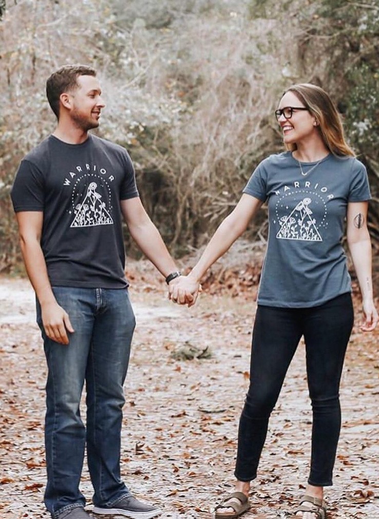 Couple holding hands wearing matching warrior t-shirts in a forest setting.