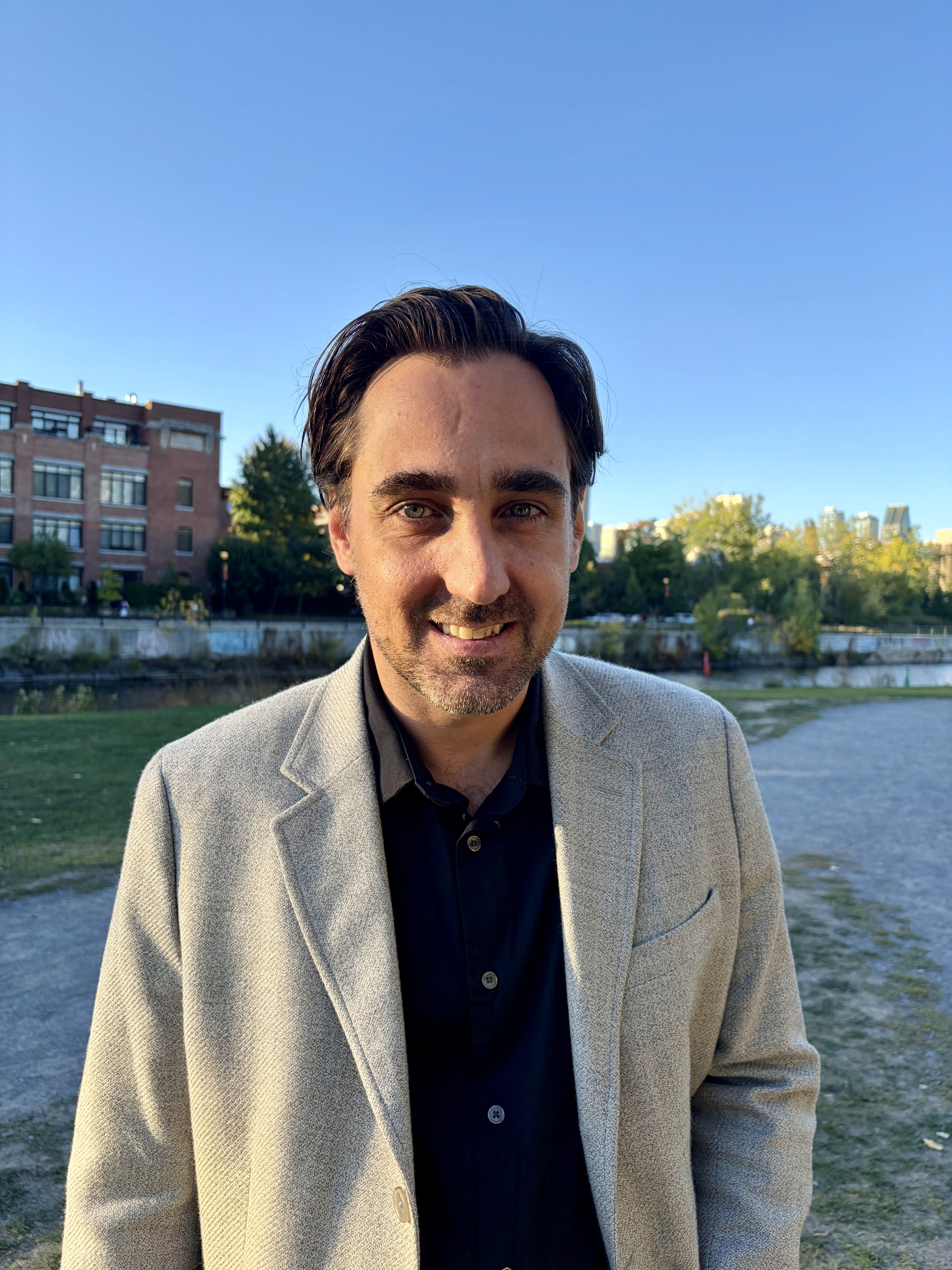 A smiling man with short hair stands with arms crossed, wearing a dark shirt against a neutral background.